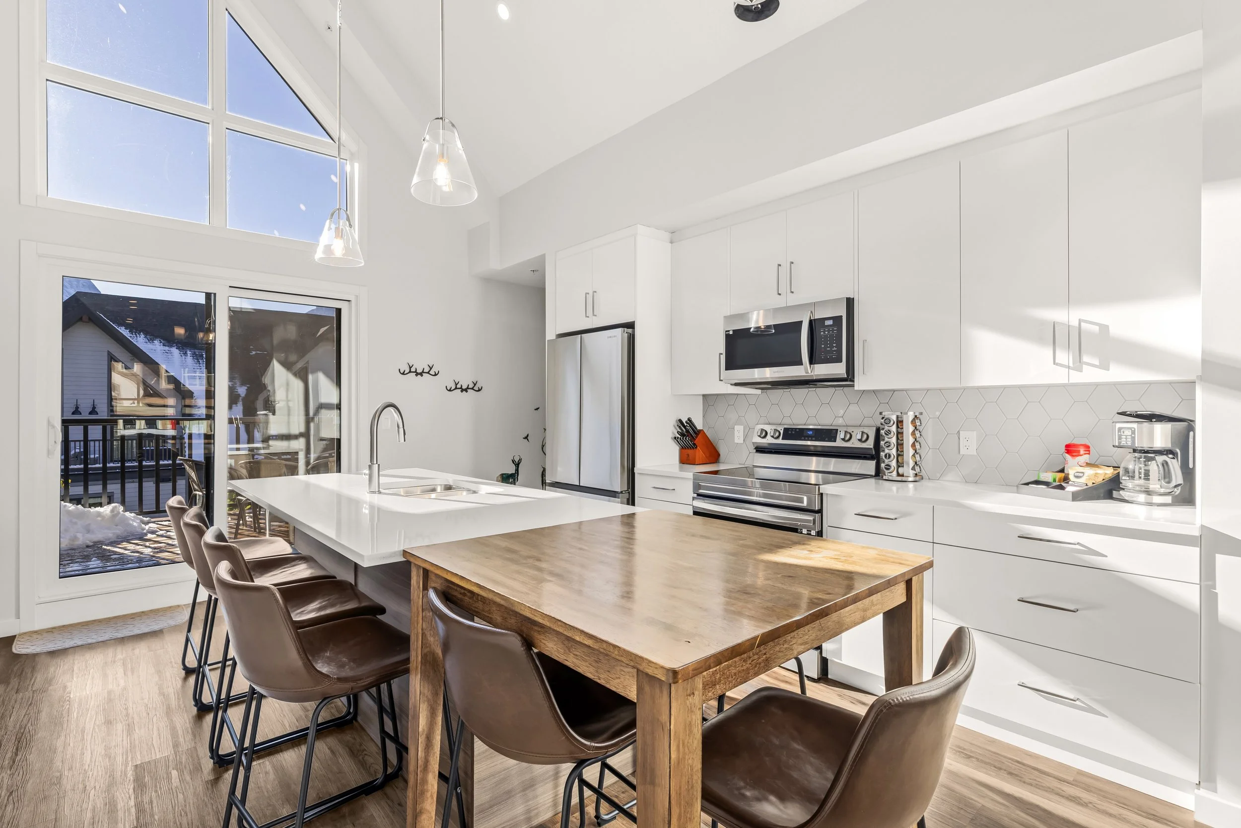 Modern kitchen with white cabinets, stainless steel appliances, a wooden dining table, and bar stools near a large sliding glass door leading to a balcony outdoor area.