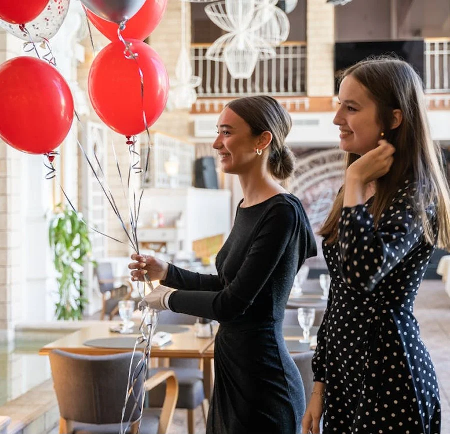 Two women at a celebration event holding red and silver balloons, smiling in a decorated indoor space.