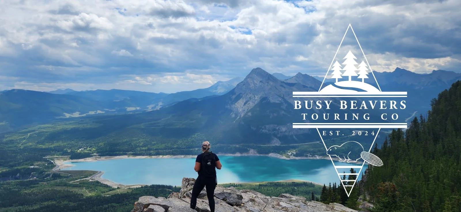 A person standing on a rock ledge overlooking a mountain lake with mountains and a cloudy sky in the background, overlaid with a logo for Busy Beavers Touring Co, featuring a triangular design with trees, a beaver, and a pinecone, and text indicating the company's name, establishment year 2024.