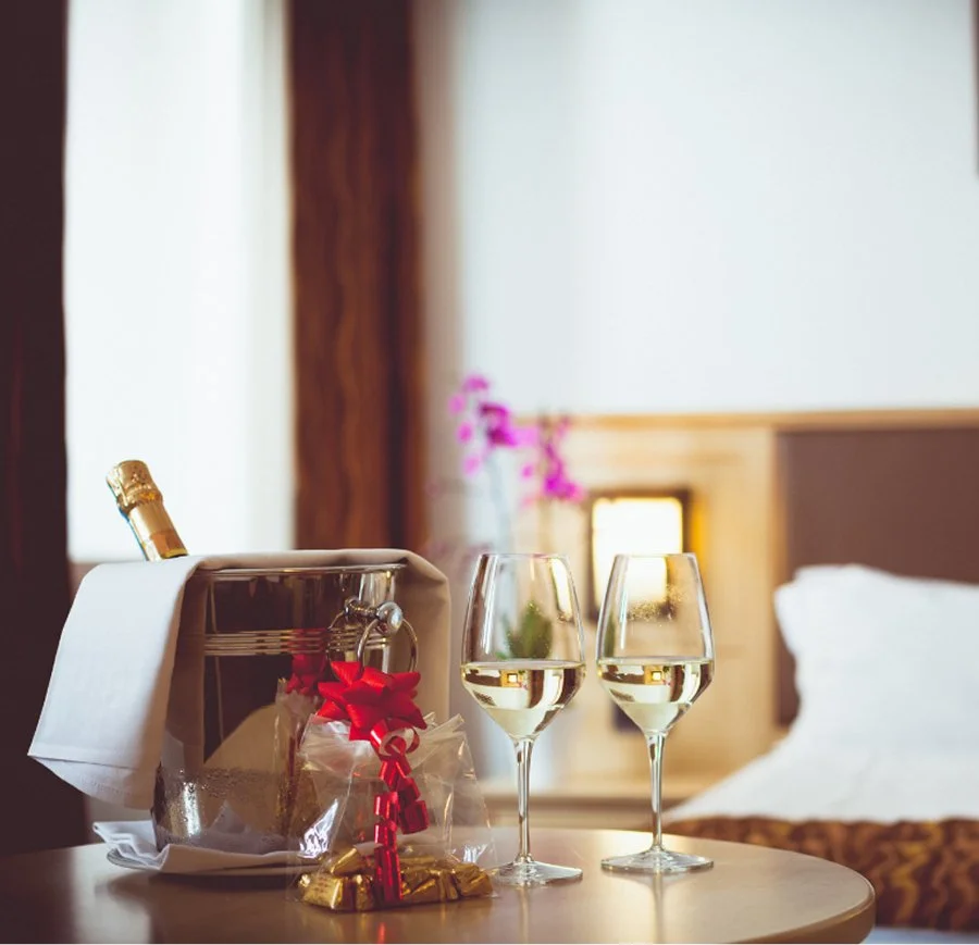 Two glasses of white wine on a nightstand in a hotel room, with a bottle of champagne in an ice bucket wrapped in a white napkin, and a small gift with a red ribbon, against a background of a bed and a blurred lamp and plants.