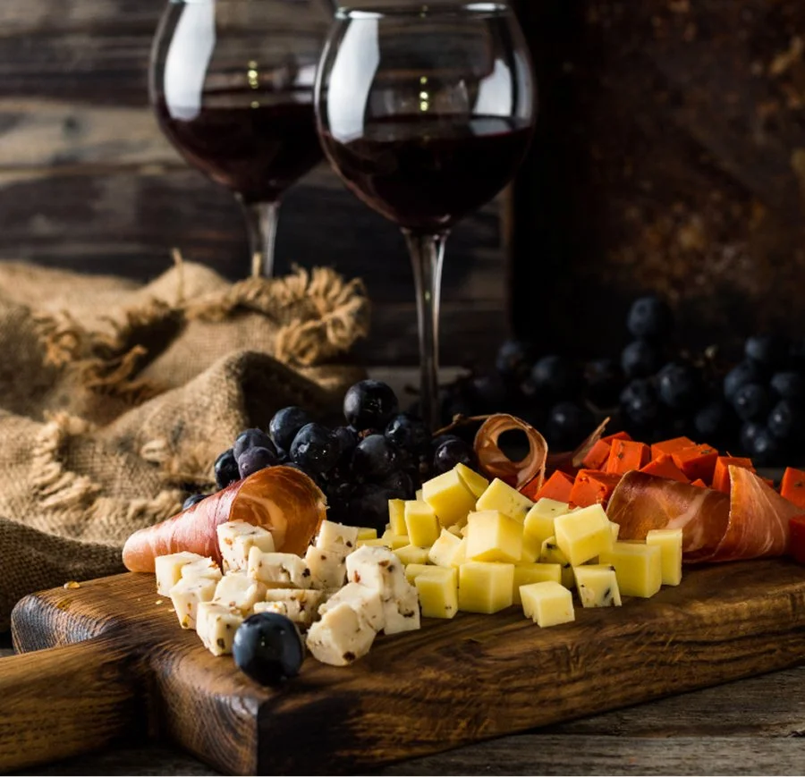 Cheese and grapes on a wooden board with glasses of red wine in the background.