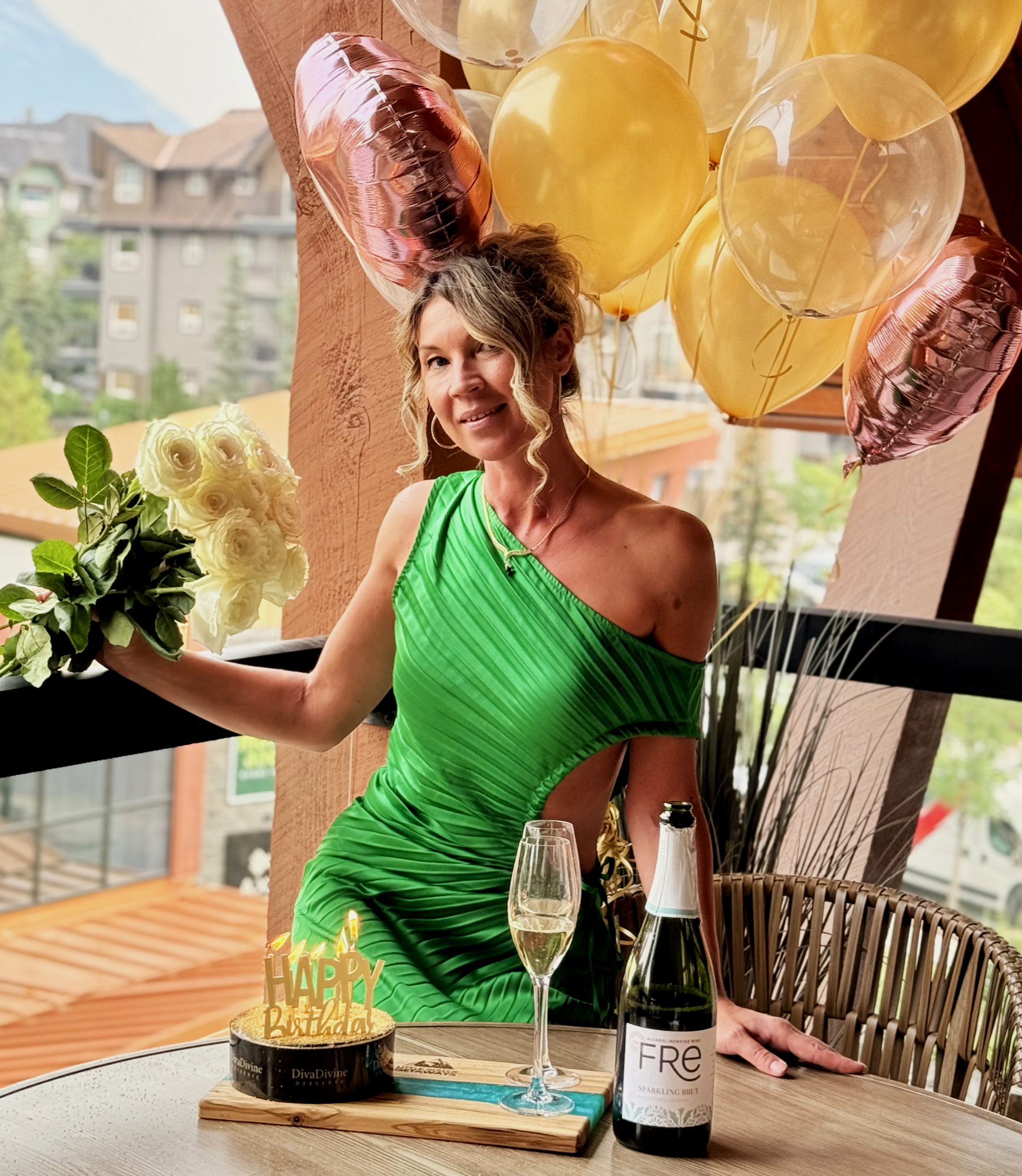 Woman in a green dress celebrating birthday with balloons, flowers, cake, champagne, and a