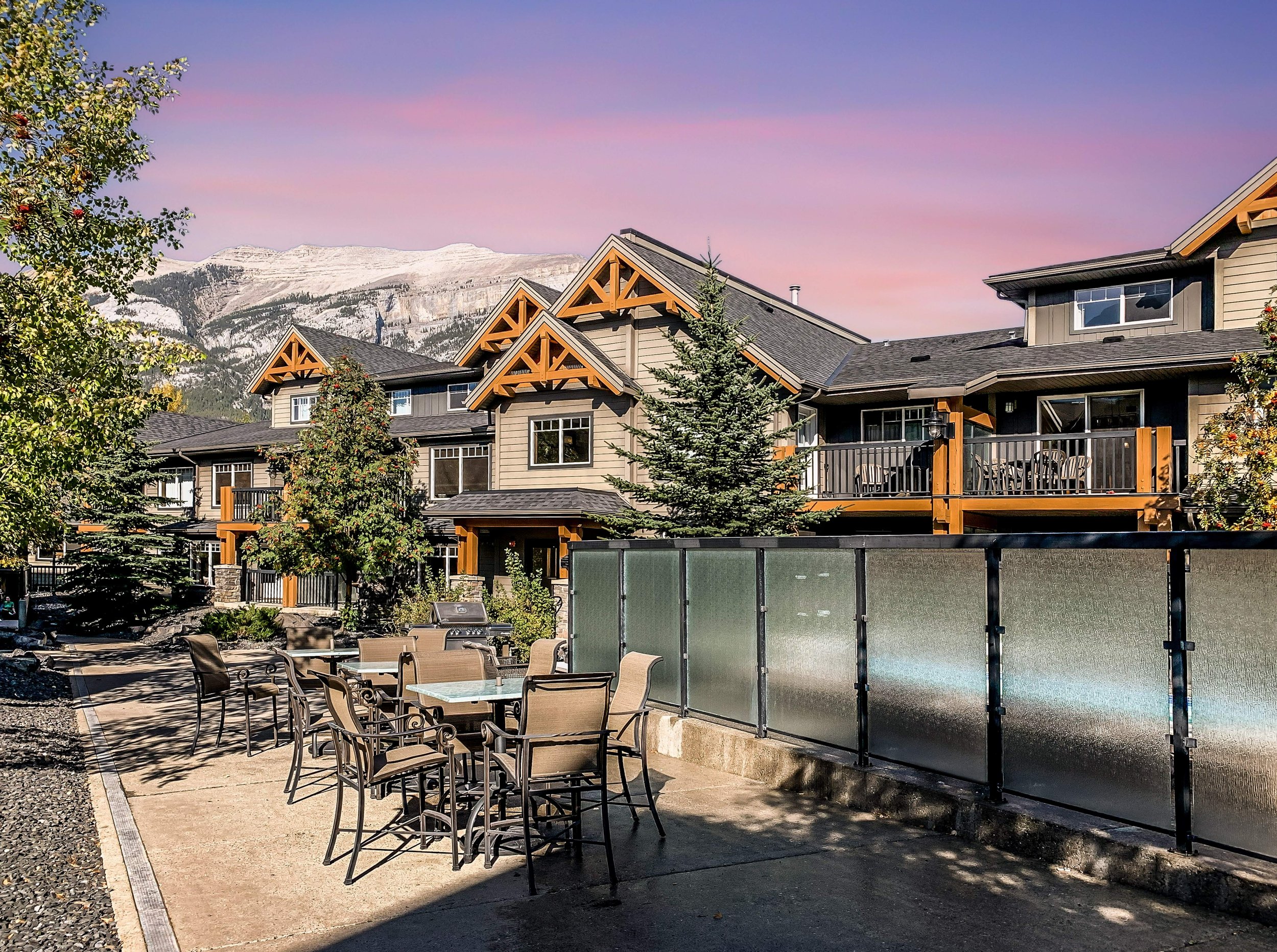 A residential backyard with a swimming pool surrounded by a stone tile deck and a frosted glass privacy fence. In the background, side view of multi-story houses with dark siding, windows, and some balconies. There are several trees with colorful autumn foliage, and a cloudy mountain range is visible in the distance under a sky with pink and purple hues.