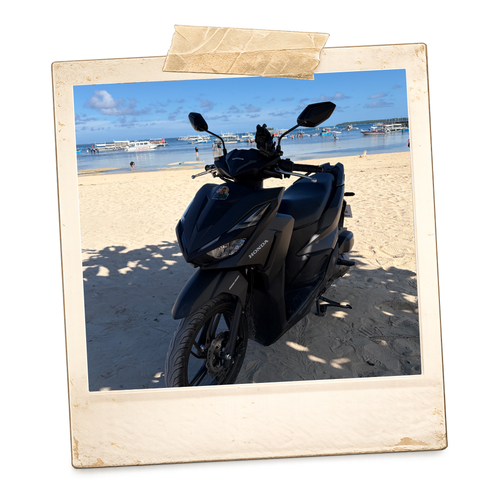 A black scooter parked on a sandy beach near the water with boats and people in the background, under a blue sky with some clouds.