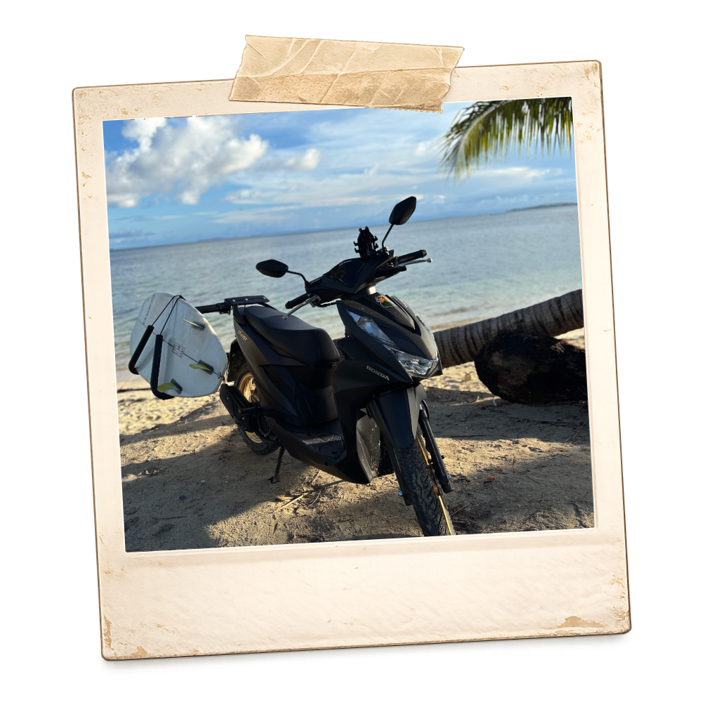 A black motorcycle with a white bag attached to the back, parked on a sandy beach near the ocean, with a leaning palm tree and blue sky with clouds in the background.