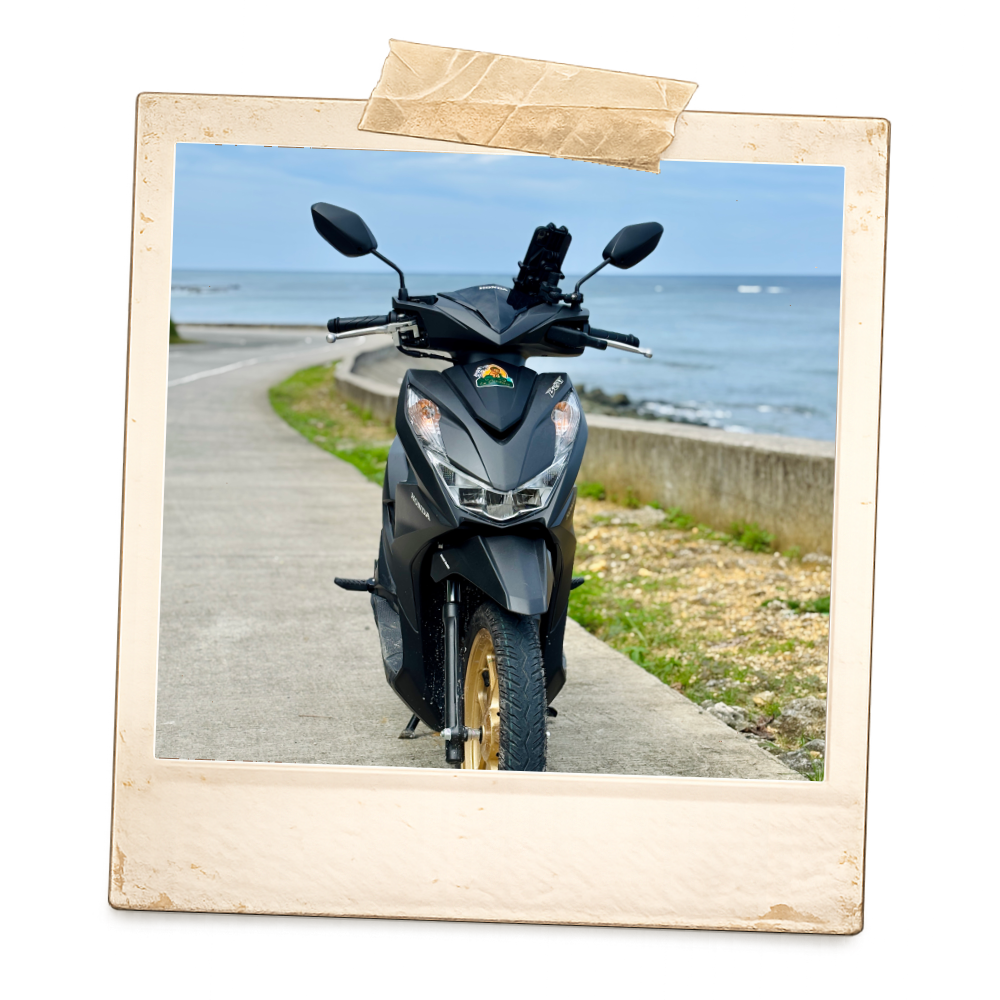 Black scooter parked on a coastal pathway with the ocean in the background and a cloudy sky.