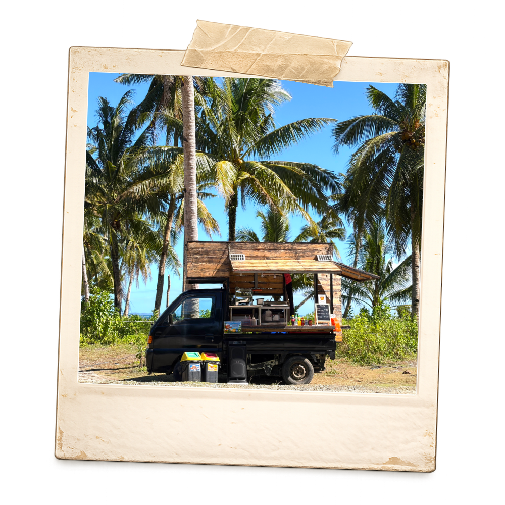 A mobile food truck resembling a mini pickup truck with a wooden serving area, parked on a tropical beach with tall palm trees and a bright blue sky in the background.
