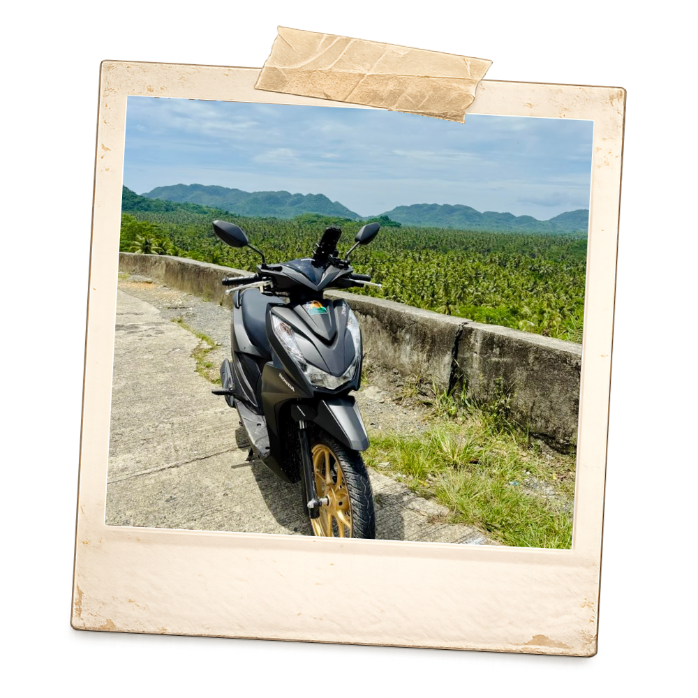 A black motorcycle parked on the side of a mountain road with green mountains and cloudy sky in the background.