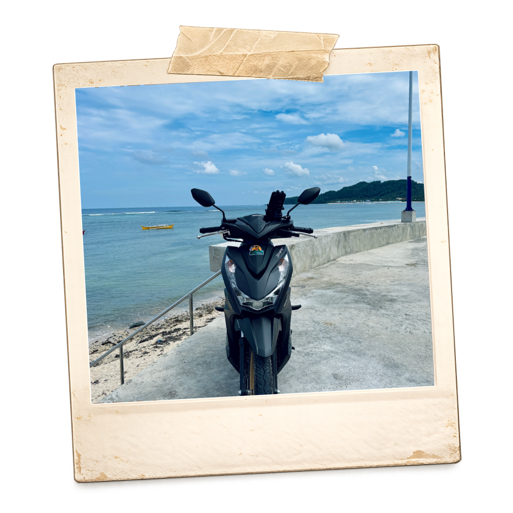 A motorcycle parked on a concrete path along a beach with a blue sky, some clouds, and a small island in the distance.
