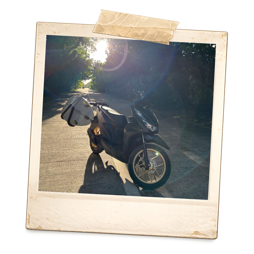 A black scooter with a side storage compartment parked on a rural road, sunlight filtering through trees, and a car visible in the distance.
