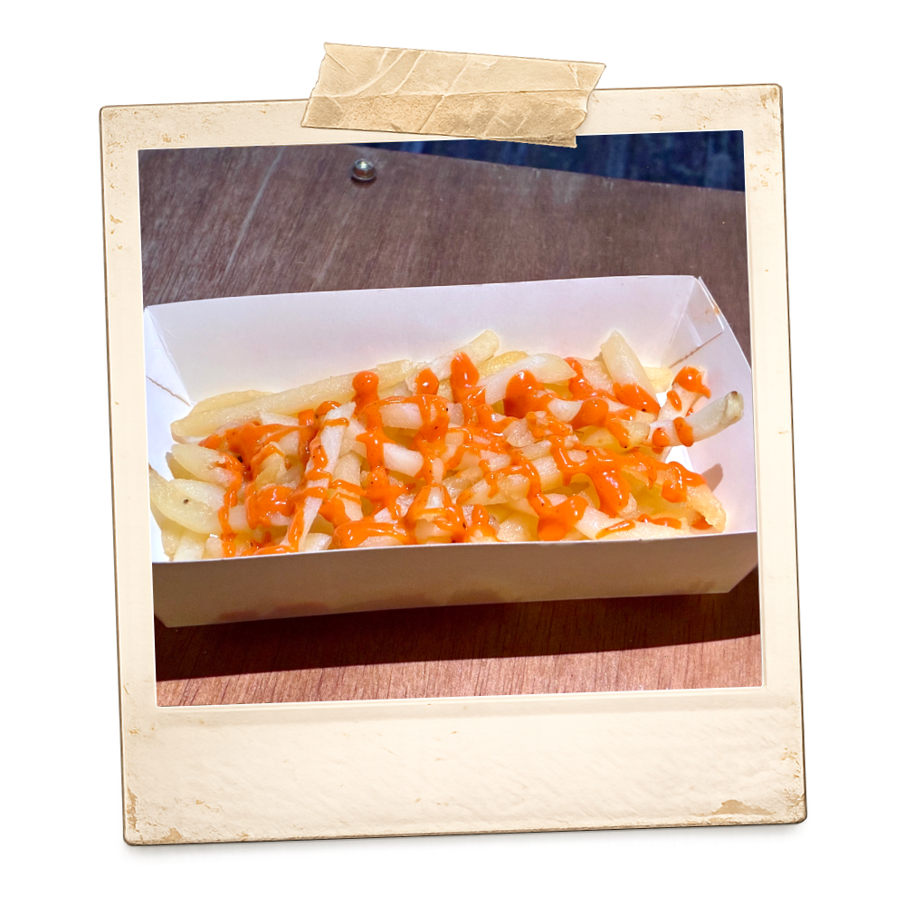 French fries topped with melted cheese and drizzled with ketchup in a white takeout container placed on a wooden surface.