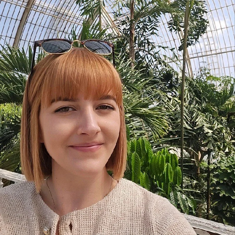 A woman with short red hair and sunglasses on her head, smiling in a greenhouse filled with lush green plants and trees.