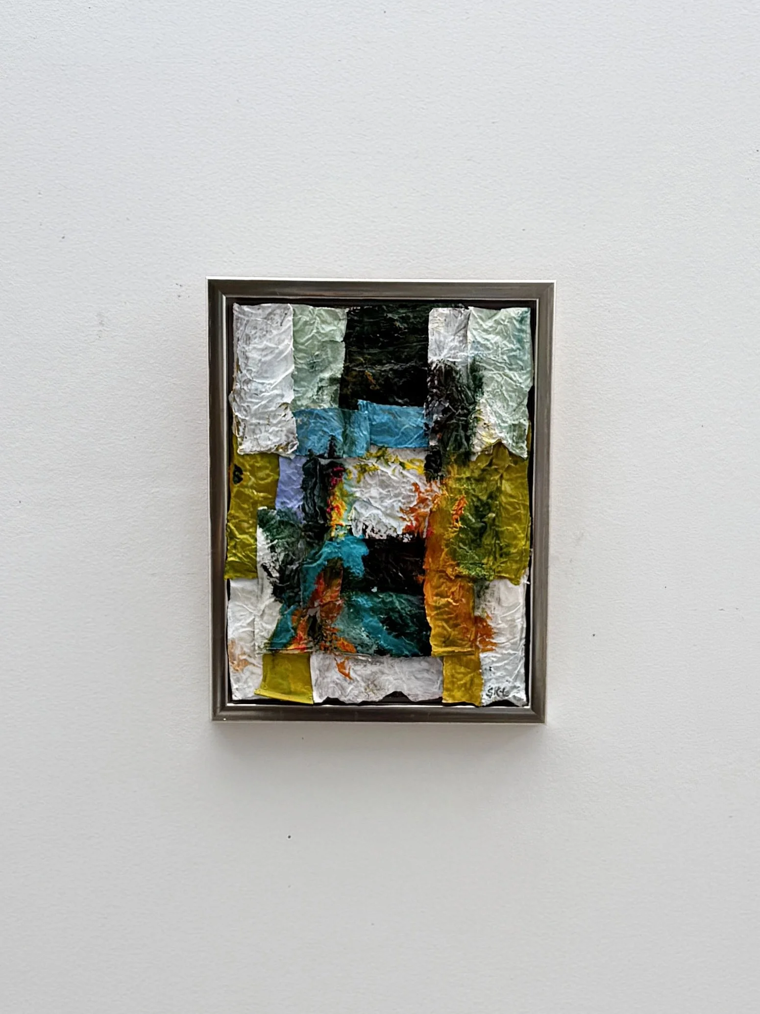 Mixed media abstract collage with torn paper, colorful textures, mounted in a silver frame on a white wall.