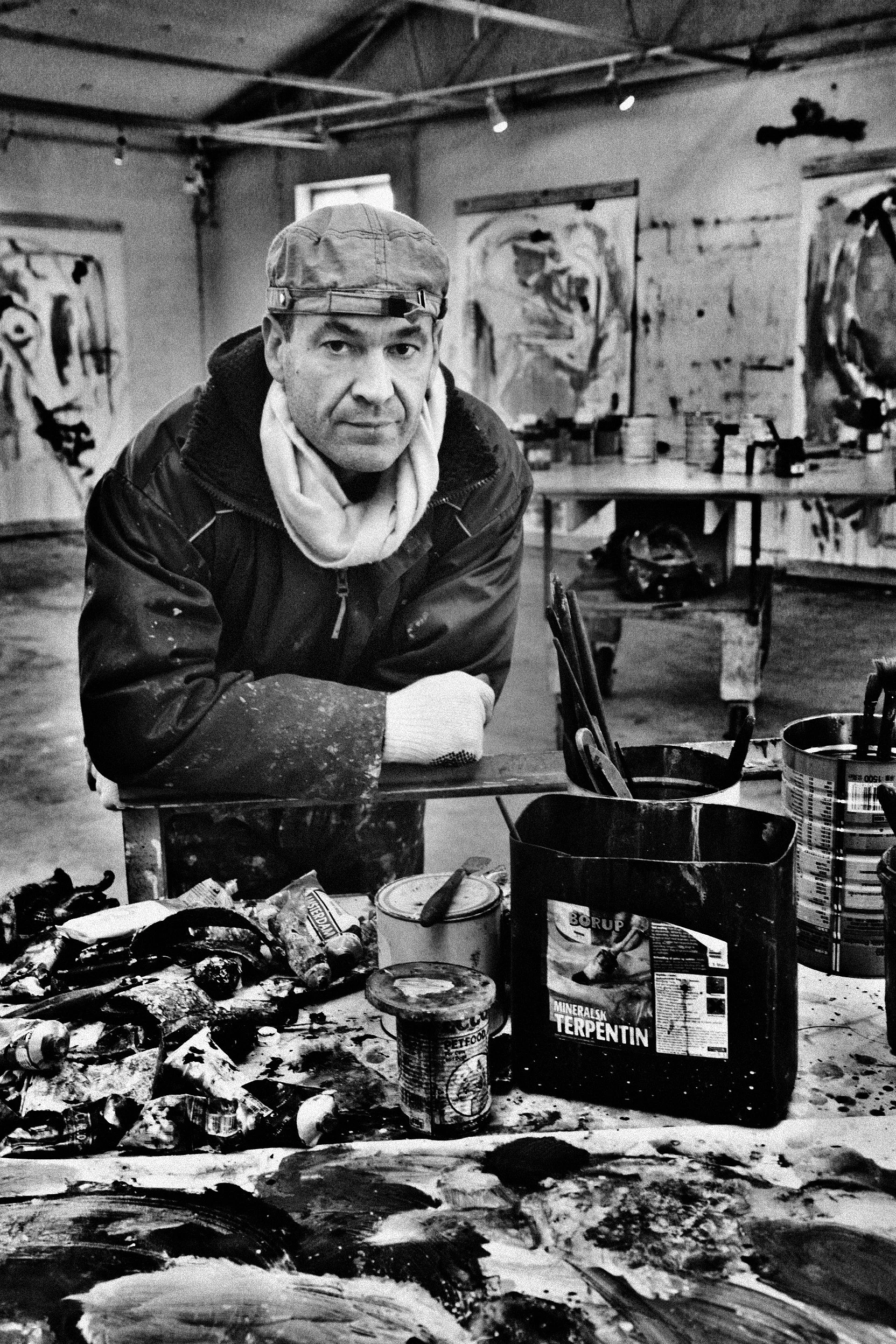 An artist in a studio with abstract paintings on the walls, leaning on a table surrounded by paint cans, brushes, and art supplies, wearing a cap, a jacket, and a scarf I Svend Kindt-Larsen I Abstract Artist I Copenhagen, Denmark
