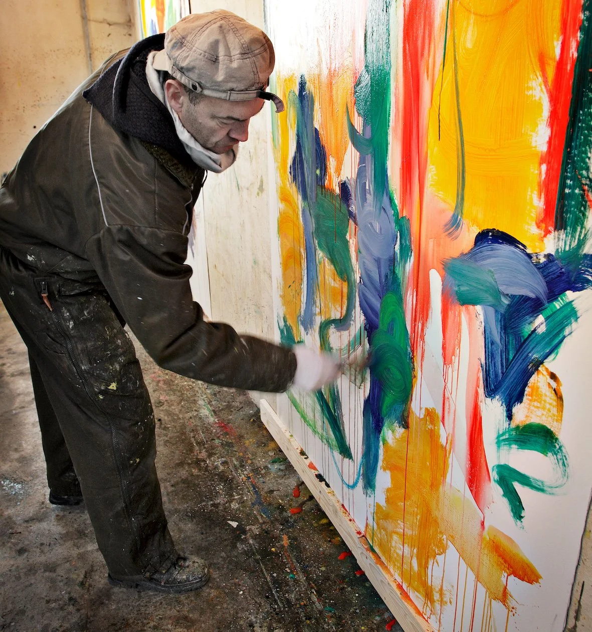 An artist in dark clothing and a beige cap creating an abstract colorful painting on a large white canvas, with splashes of green, blue, yellow, orange, and red paint I Svend Kindt-Larsen I Abstract Artist I Copenhagen, Denmark