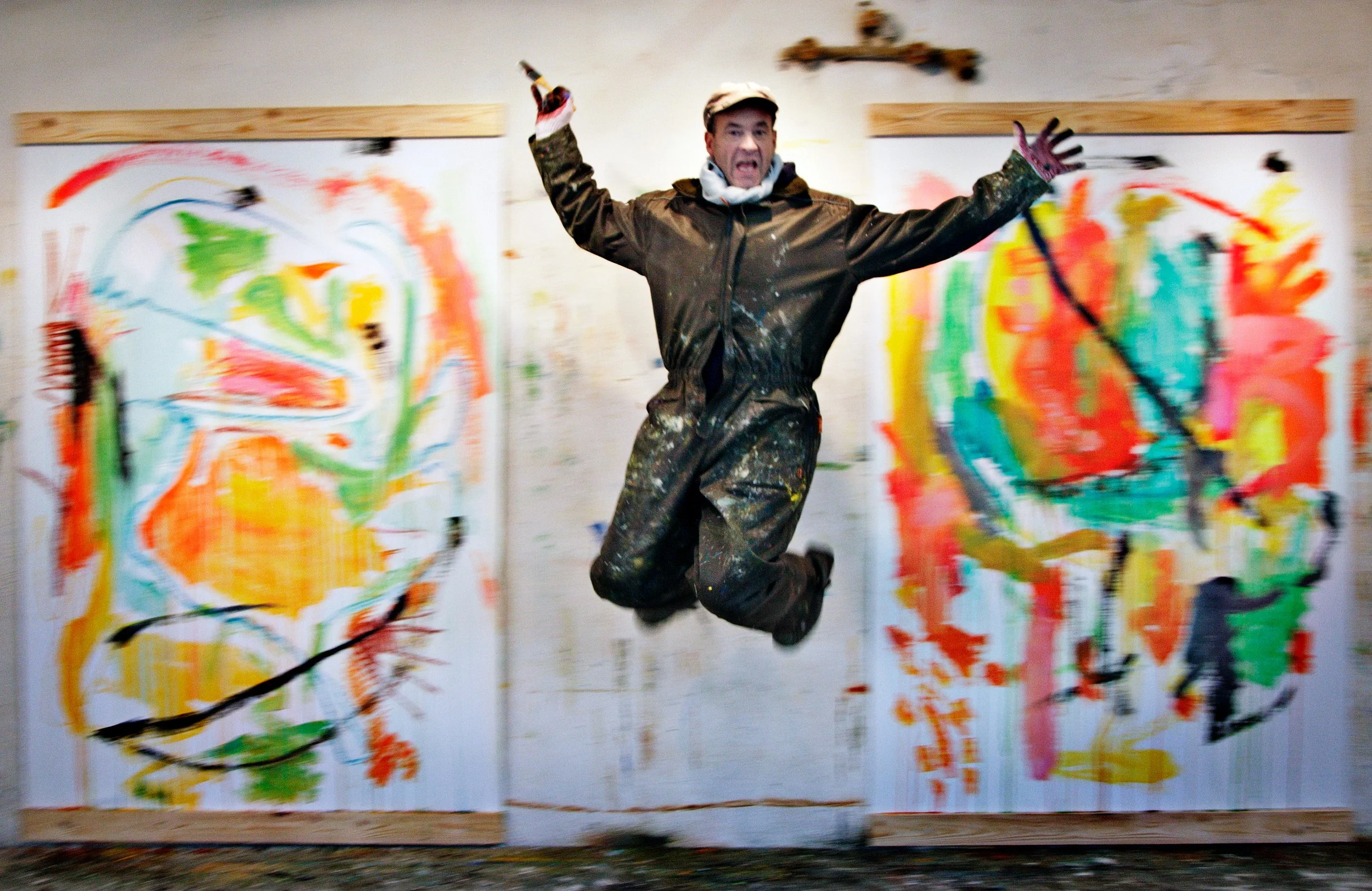 A man in a paint-stained jumpsuit and cap is jumping in front of two large, colorful abstract paintings on the wall I Svend Kindt-Larsen I Abstract Artist I Copenhagen, Denmark
