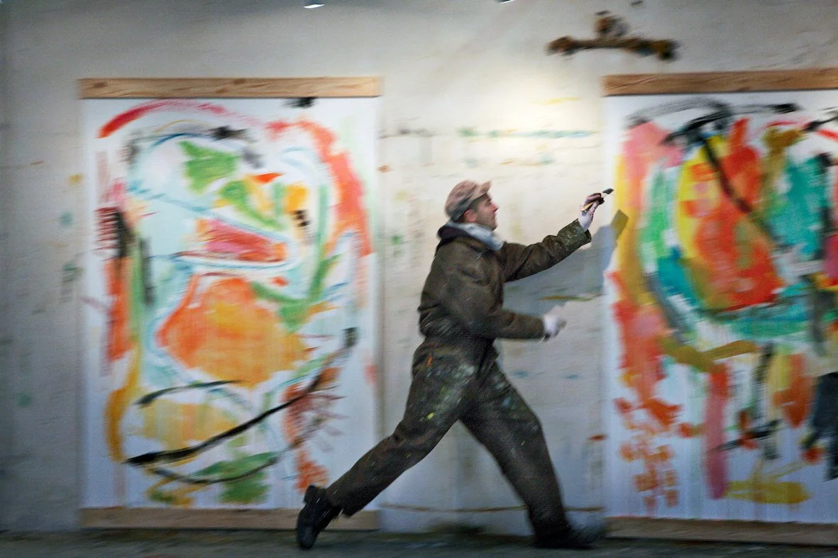 A person in dark clothing with paint splatters on it is running while holding a paintbrush and a palette in front of two colorful abstract paintings on a wall I Svend Kindt-Larsen I Abstract Artist I Copenhagen, Denmark