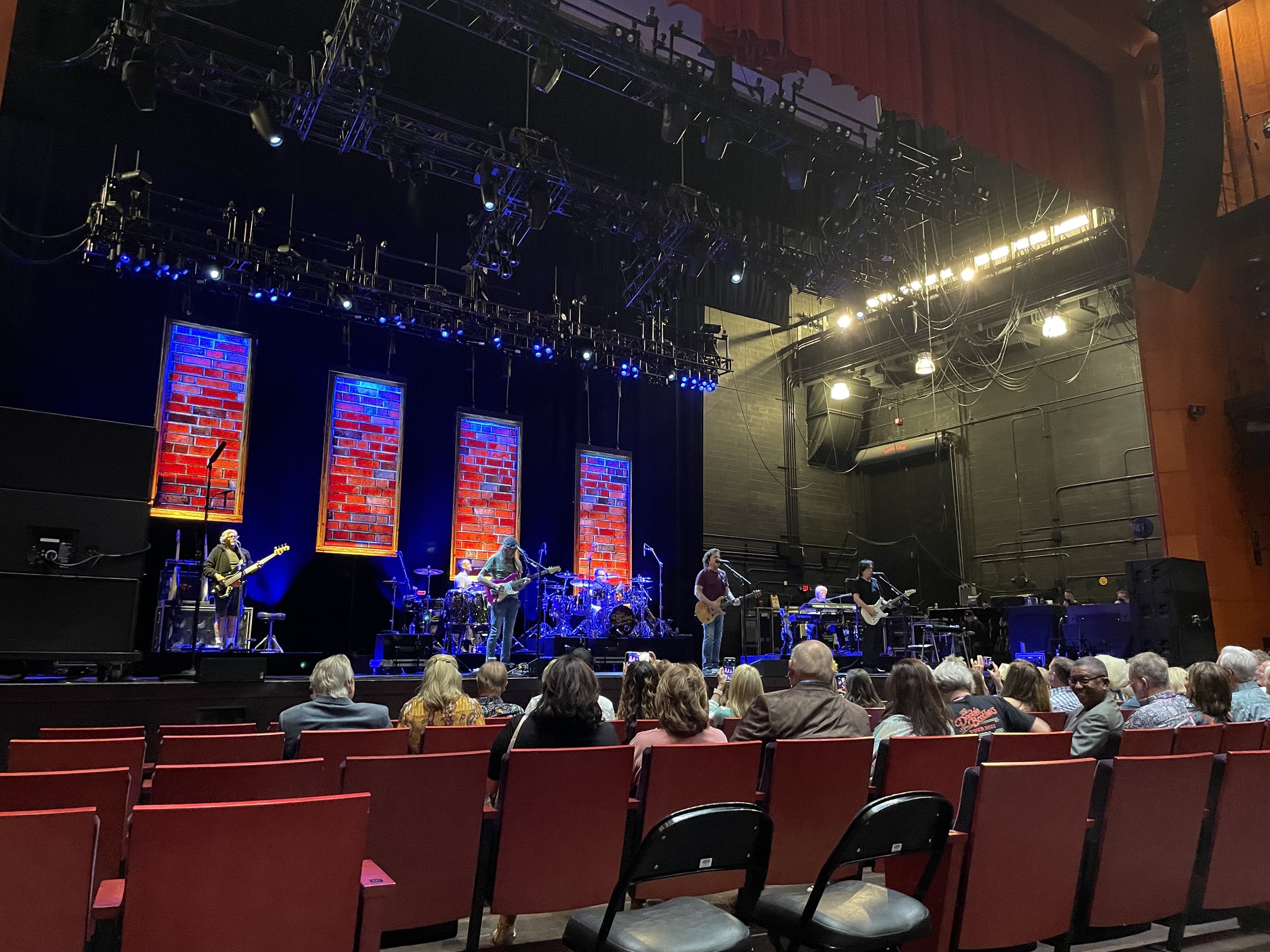A band performing on stage with colorful backdrops and an audience seated in front of the stage in a theater.