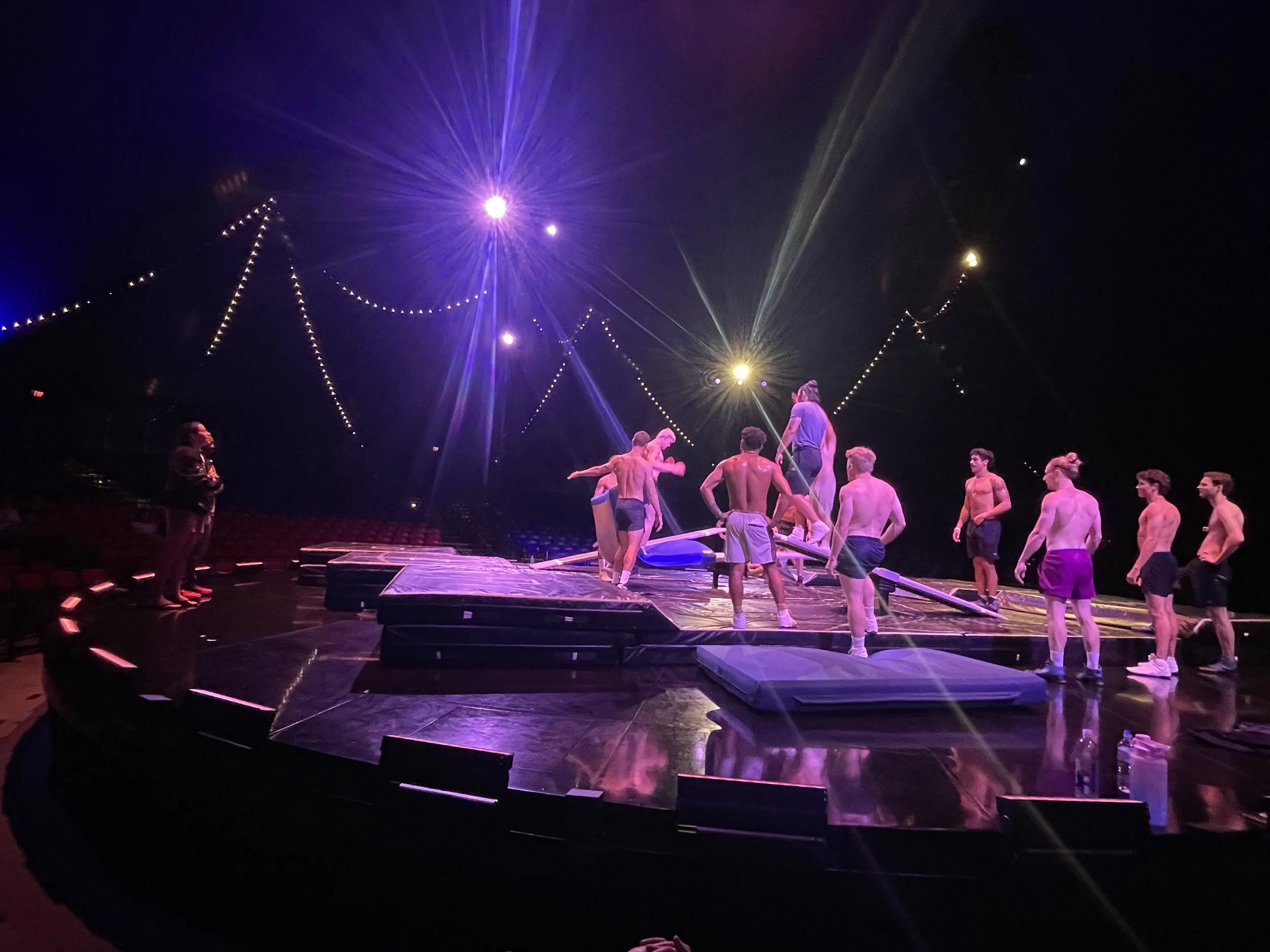 Circus performers practicing on a teeterboard under purple and yellow stage lights.