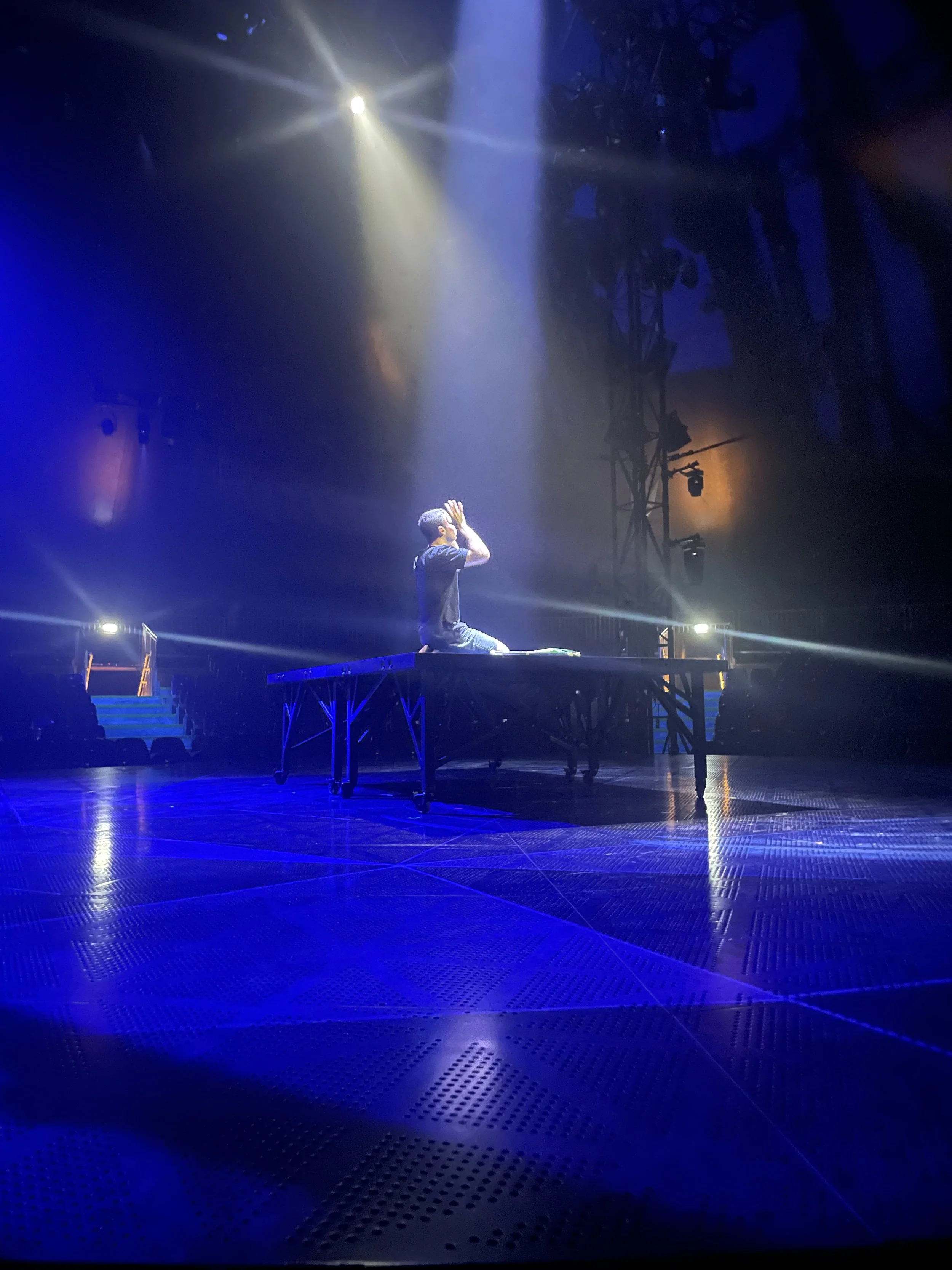 A person kneeling on a stage under a spotlight, with their hands cupped in front of their face, in a dark theater venue with colorful lighting and equipment in the background.