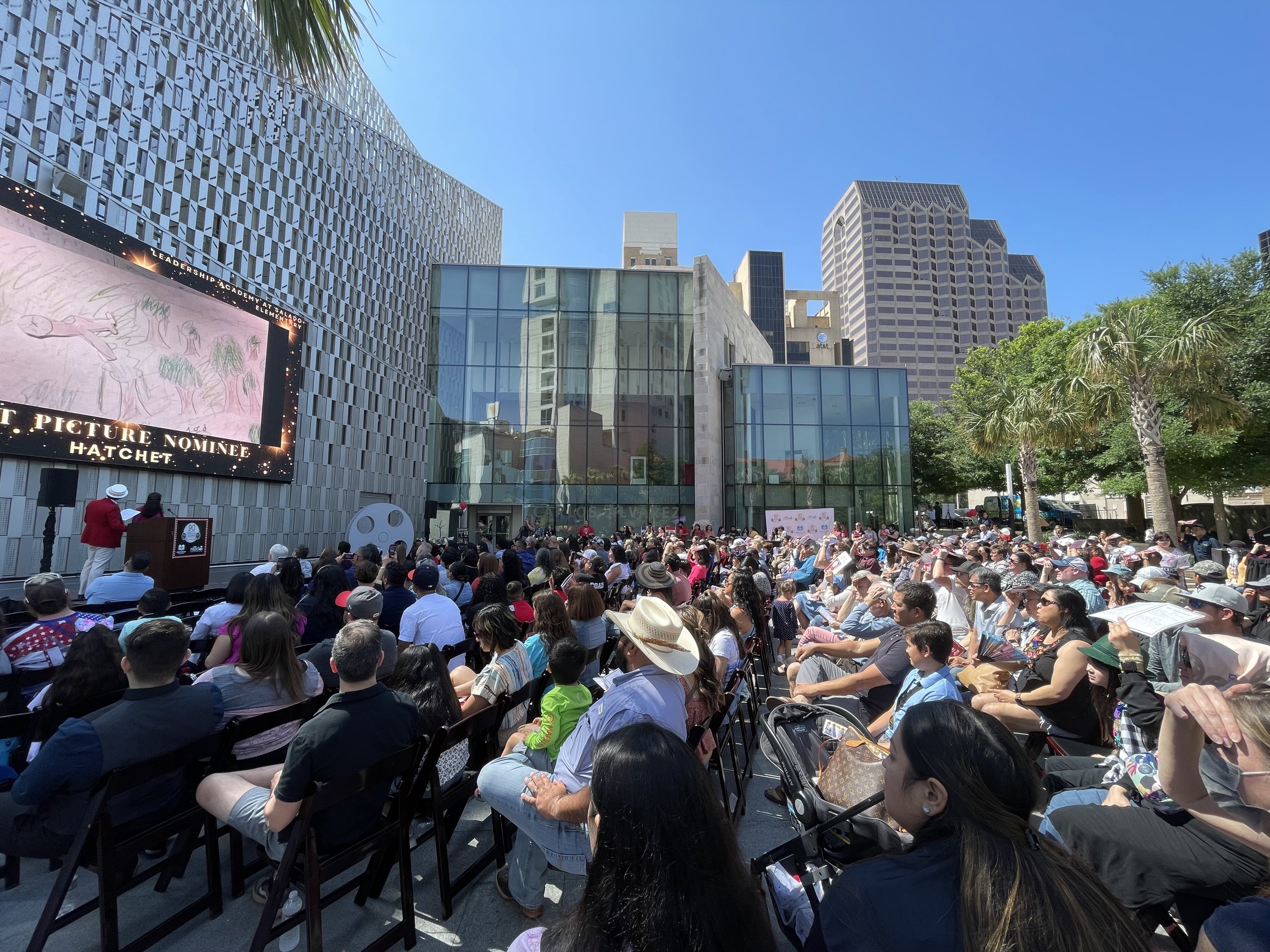 A large outdoor event in a city with a crowd of people seated facing a stage outside the Tobin Center, with seating arranged in front of a modern glass building. The scene is on a sunny day with blue skies and palm trees.