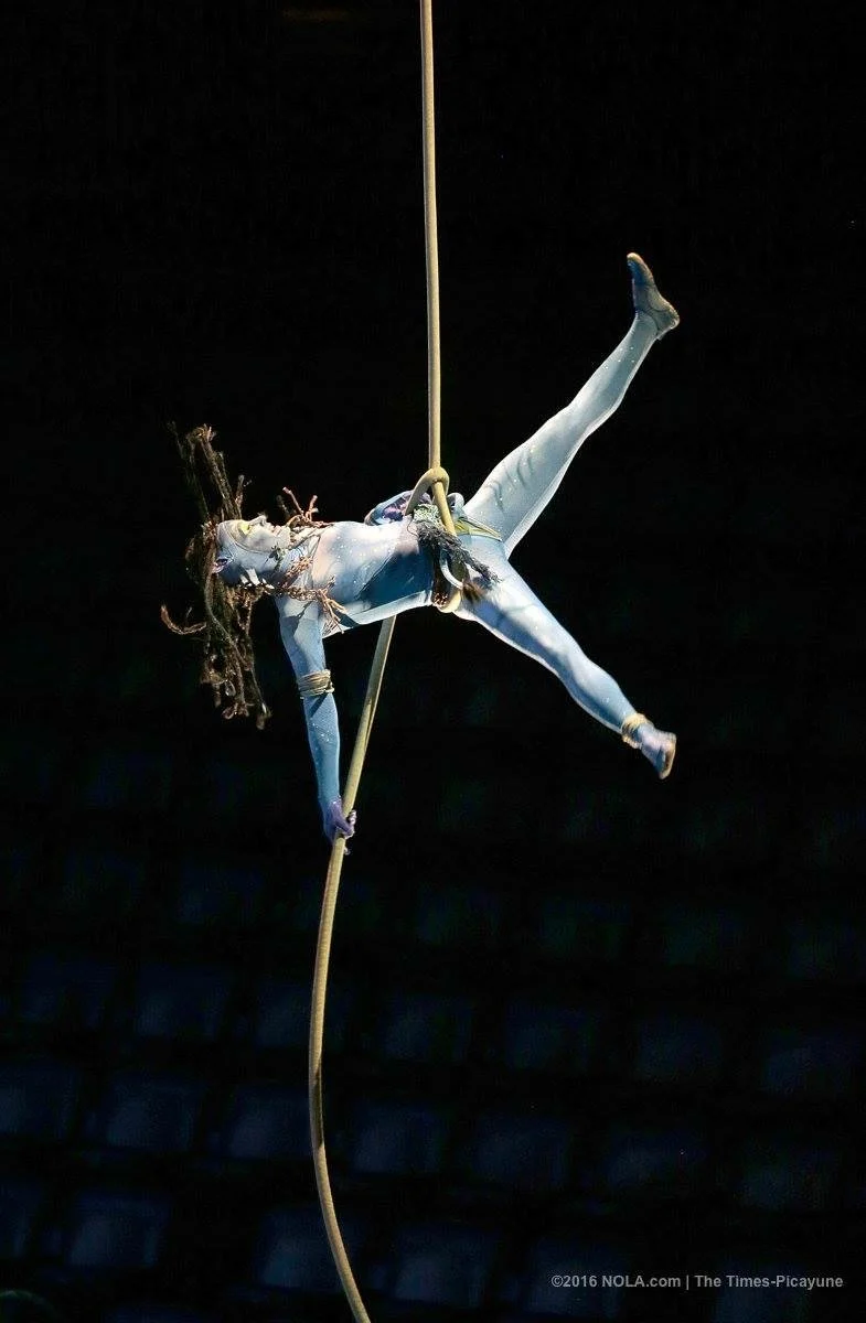 Aerial performer dressed as a character from Avatar, with blue skin and tribal makeup, hanging from a rope during a stage performance in an indoor arena against a dark background.