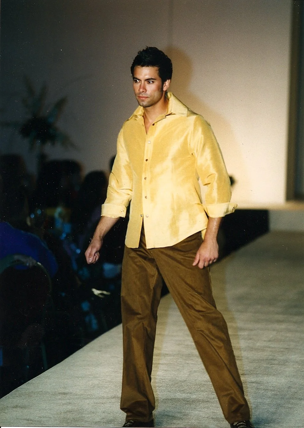 Male model walking on a runway during a fashion show, wearing a shiny yellow shirt and brown pants.