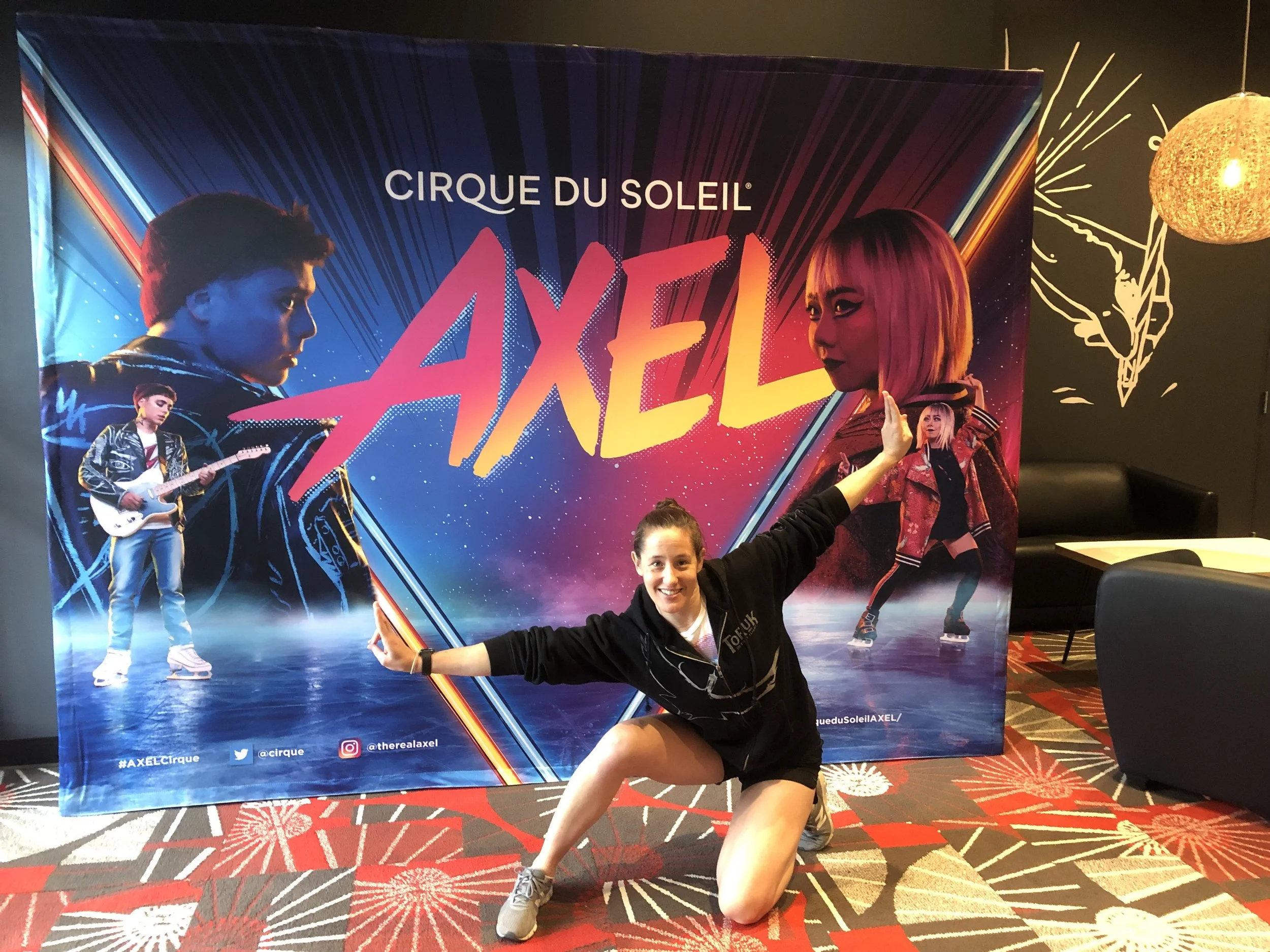 A young woman kneeling and smiling in front of a burnout poster for Cirque du Soleil's AXEL show, with one arm raised and the other pointing at the poster.