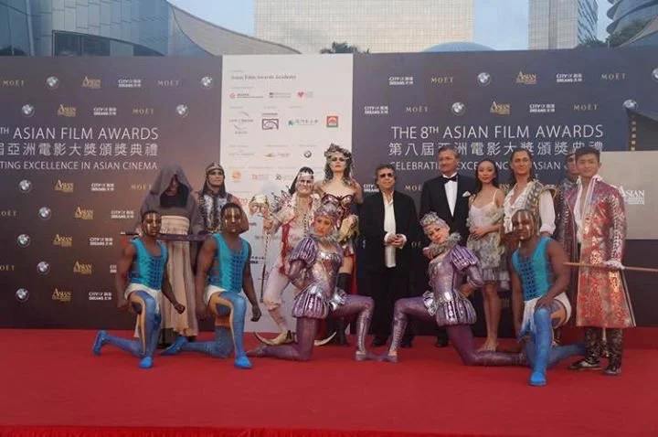 Group of people on red carpet at the 8th Asian Film Awards in Macau, posing for photograph, with some in costumes and glamorous attire, backdrop with event information.