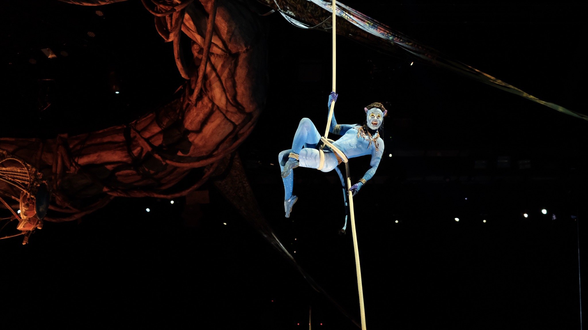 A performer dressed as a character from Avatar, with blue skin and tribal makeup, hanging from a rope during a stage performance in an indoor arena, with a large, intricately designed piece of set in the background.