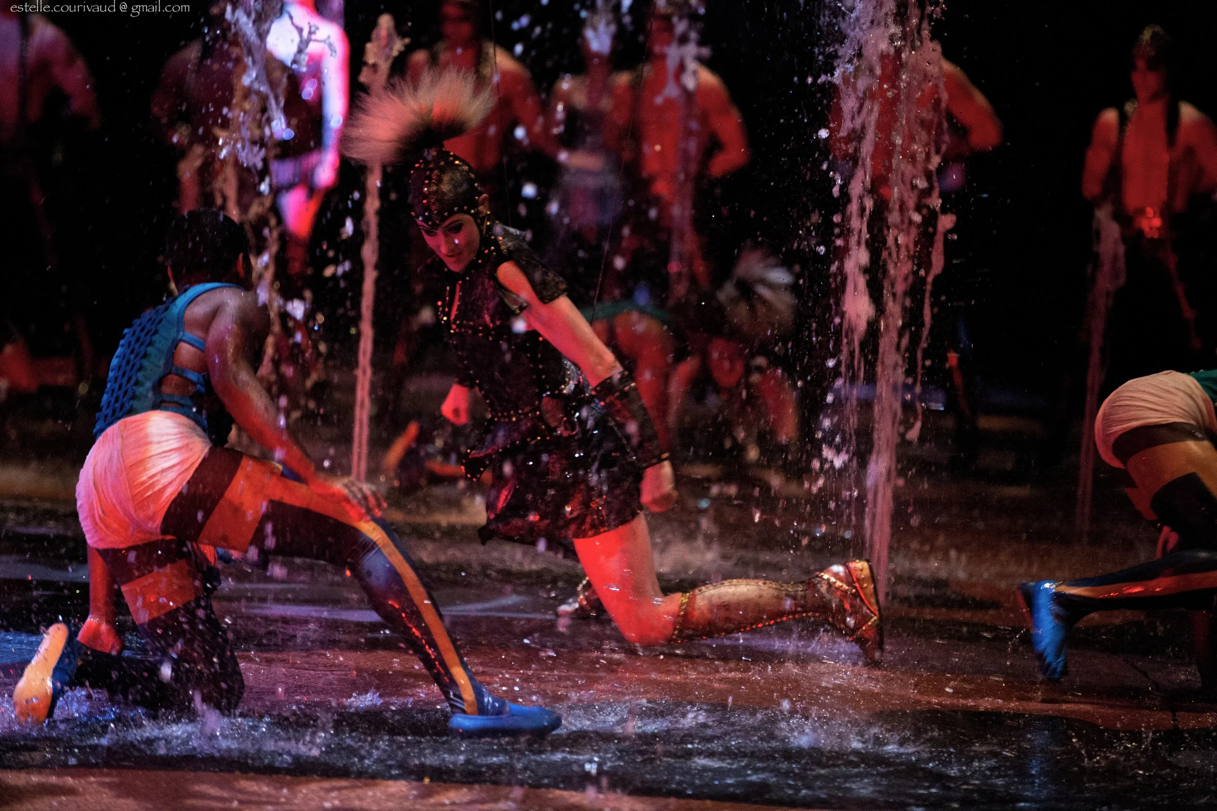 A performer dressed in a black costume performs in a water-themed theatrical stage, surrounded by water sprays and other performers in colorful costumes.