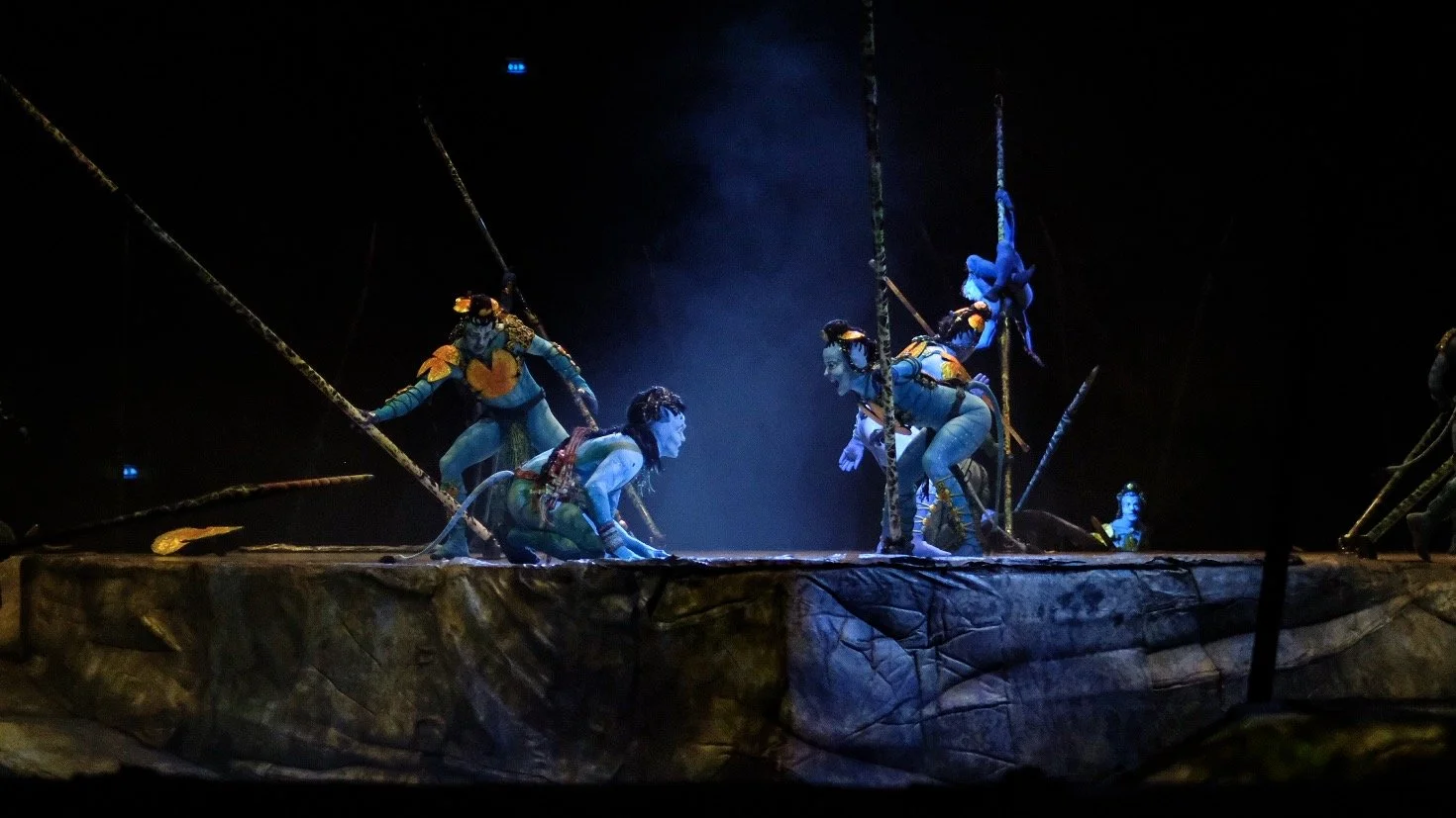 Performers dressed as characters from Avatar on a stage with a rocky set and suspended poles, mid-performance, under blue lighting.