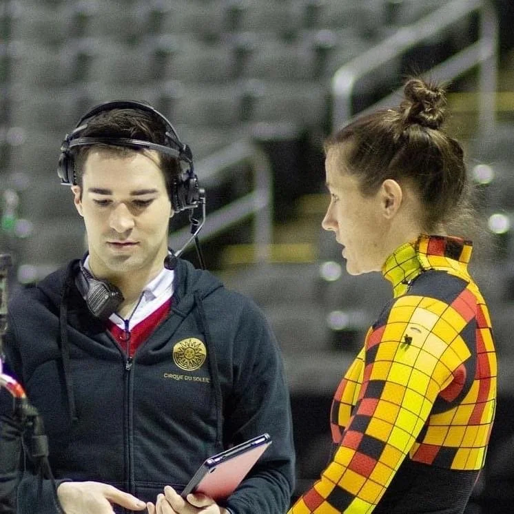 Two people in a conversation, one man with dark hair and a headset, and one woman with her hair in a bun, wearing a yellow and red checkered costume, in an indoor arena with gray seats in the background.