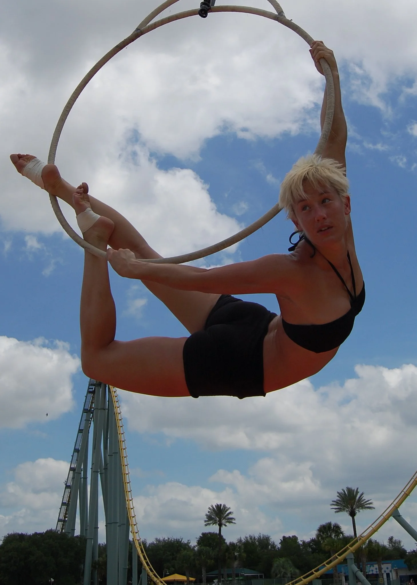 A woman with blonde hair in black swimsuit performing an aerial hoop skill outdoors at an amusement park with roller coaster and palm trees in the background.