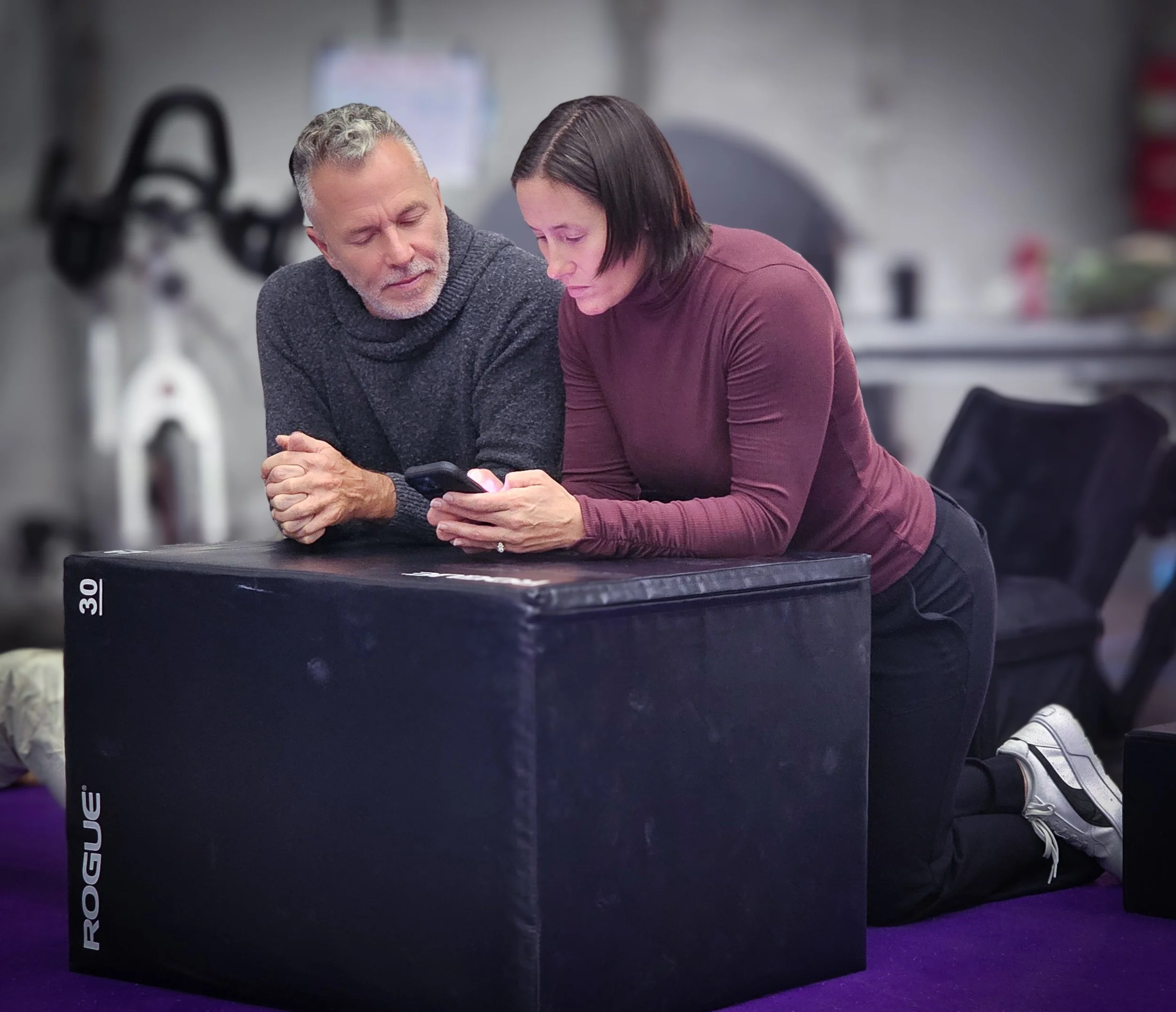 A man and woman kneeling on a purple mat in a gym, looking at a smartphone together. The woman is wearing a maroon long-sleeved top and black pants, the man is wearing a dark gray sweater. There is a black Rogue workout box in front of them and gym equipment in the background.