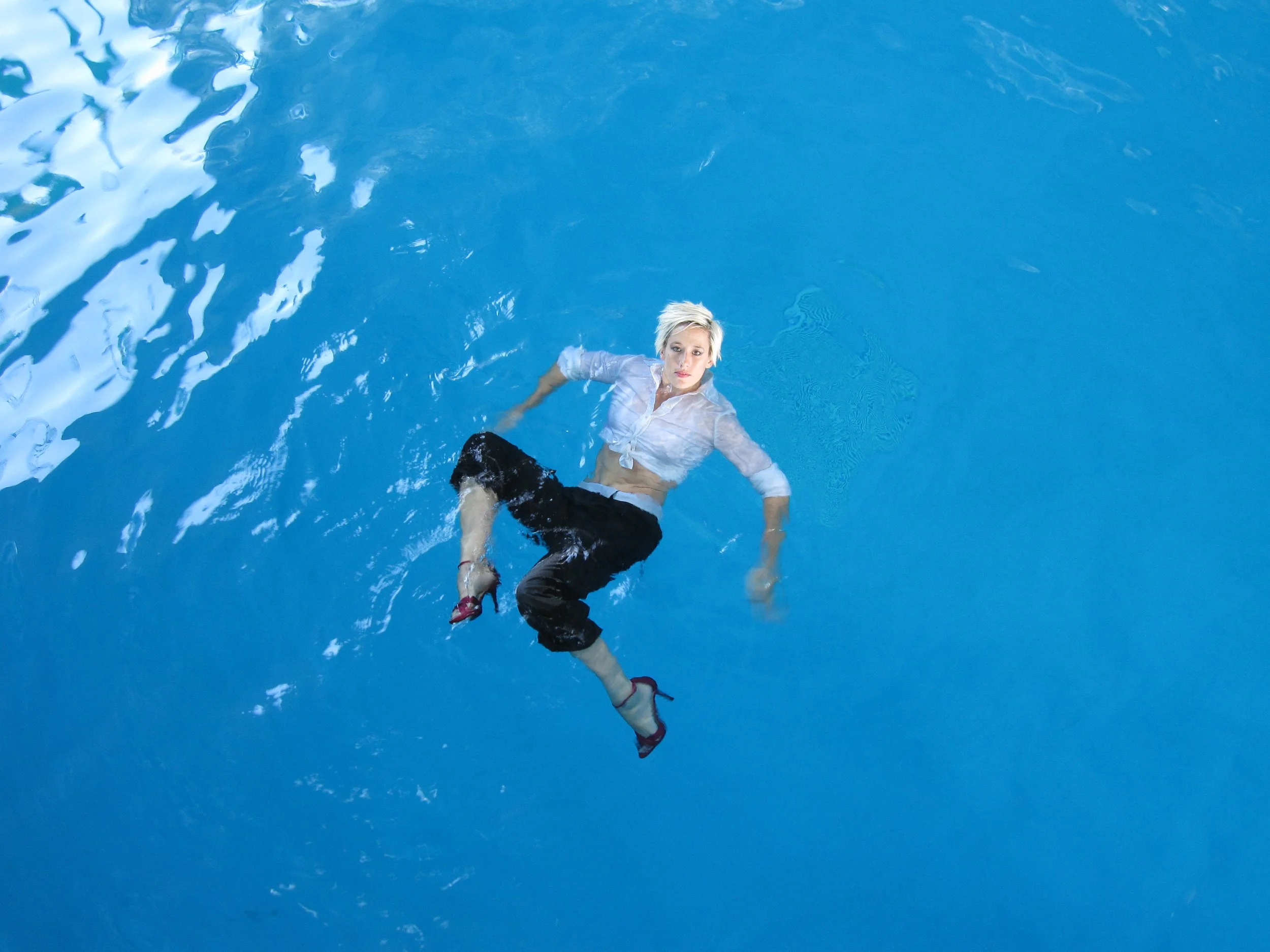 A woman with short blonde hair wearing a white shirt, black pants, and pink high heels floating on her back in a swimming pool.