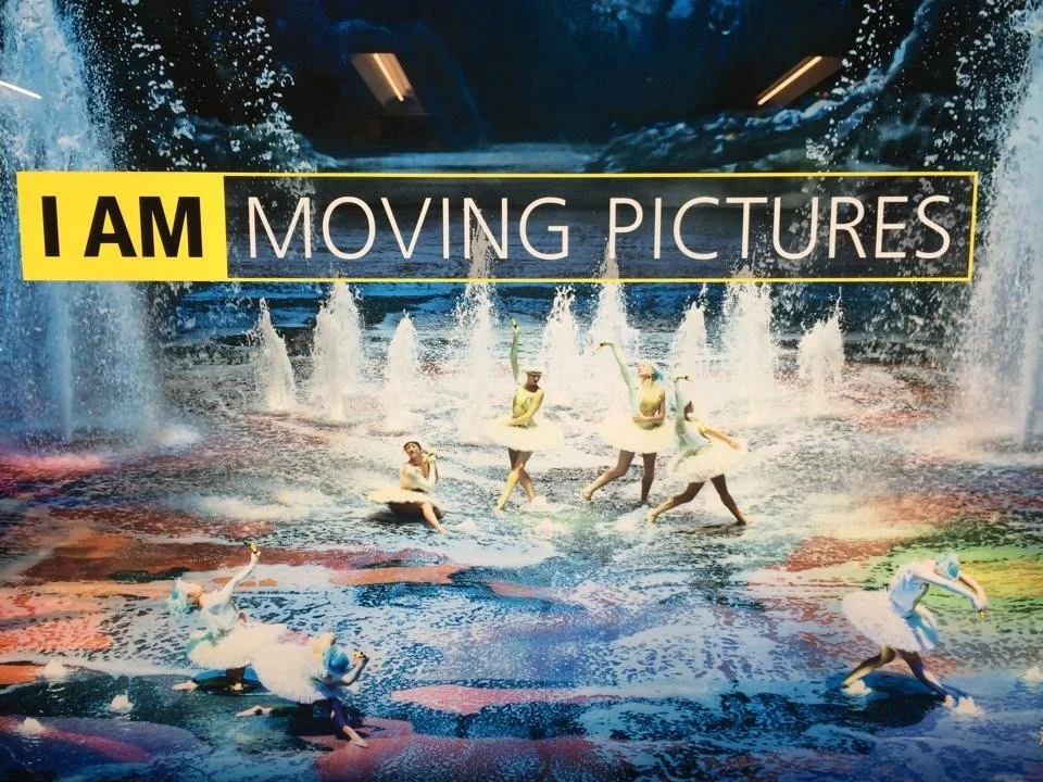 Colorful Nikon advertising campaign poster with the text 'I AM MOVING PICTURES' and images of dancers in swan-like ballet costumes posing on stage with a water fountain backdrop at The House of Dancing Water in Macau.