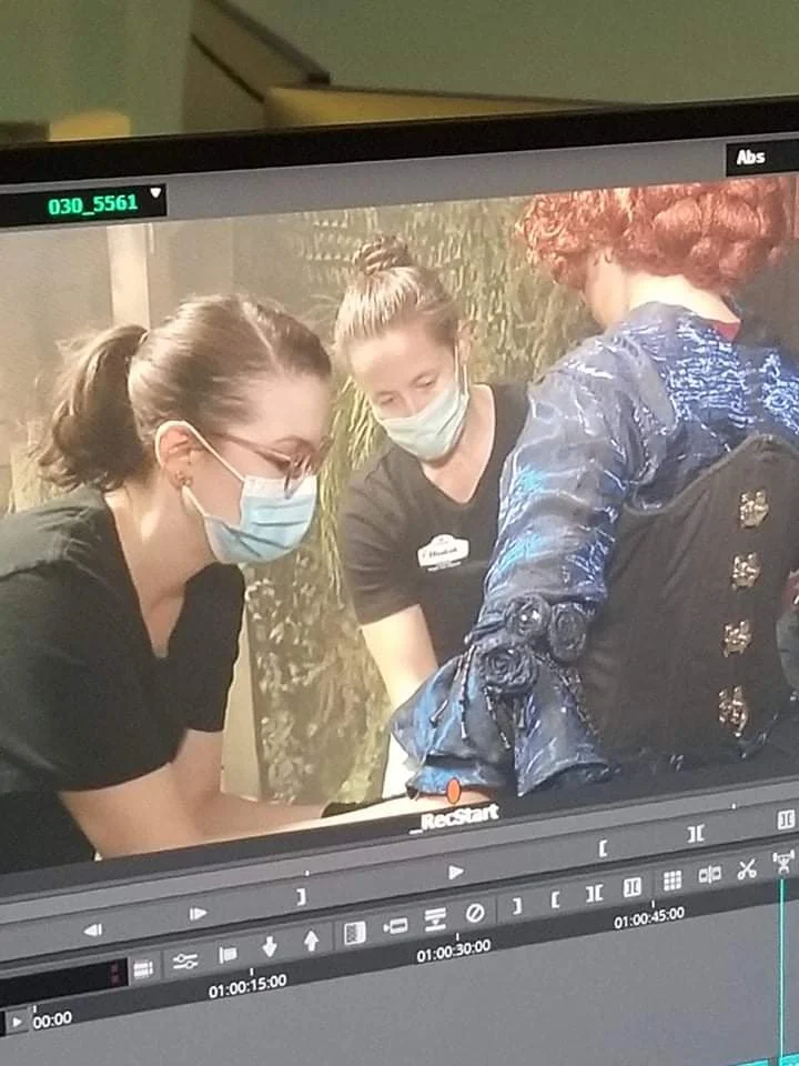 Three women wearing face masks and black clothing are looking down at something out of view in front of them. Two of them have buns and one has curly red hair, and they are adjusting a costume on a performer. 