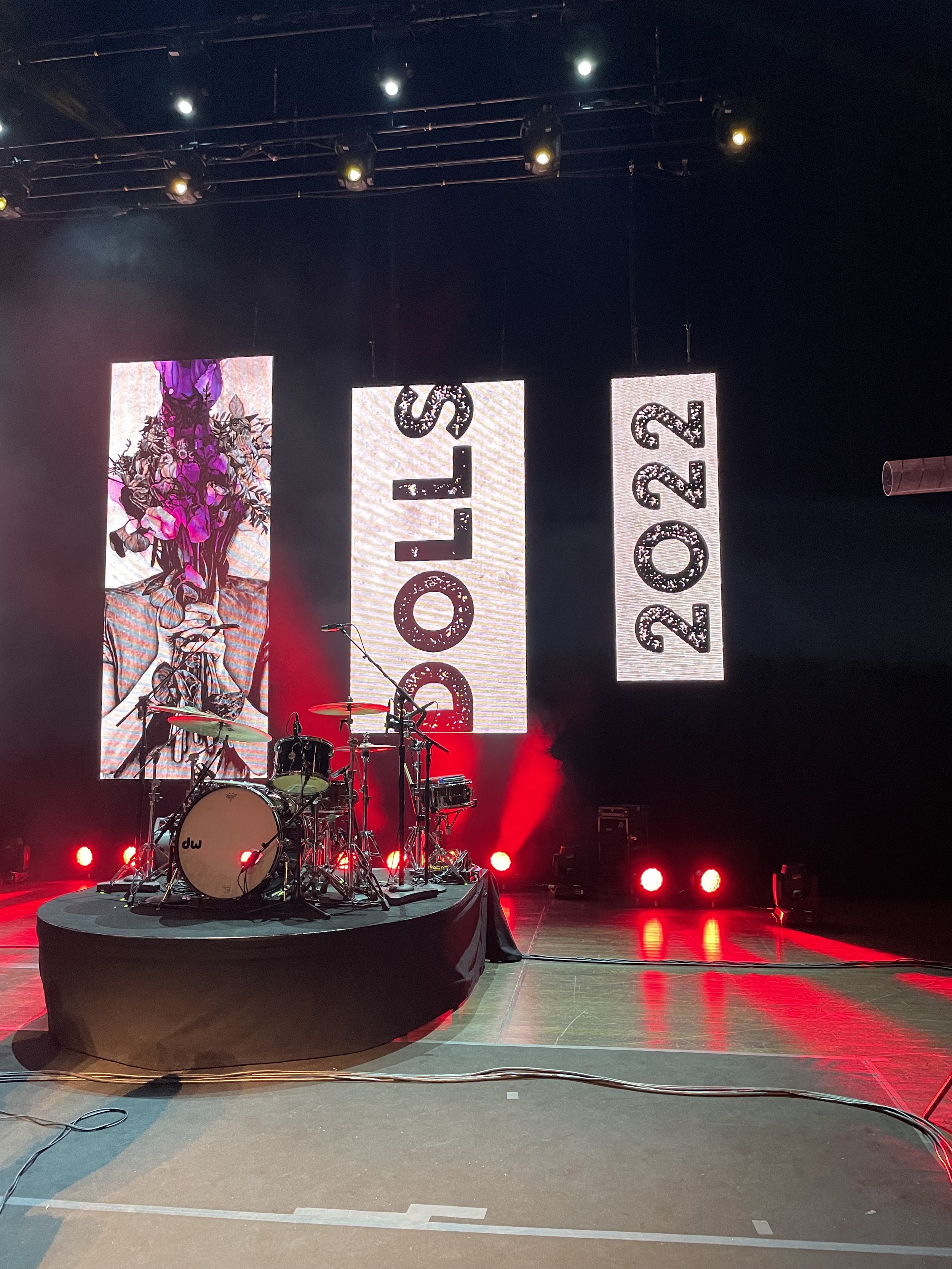 Stage with drum set on a black platform, illuminated by red lights, with three large vertical screens displaying 'DOLLS 2022' and an artistic image of a face with flowers.