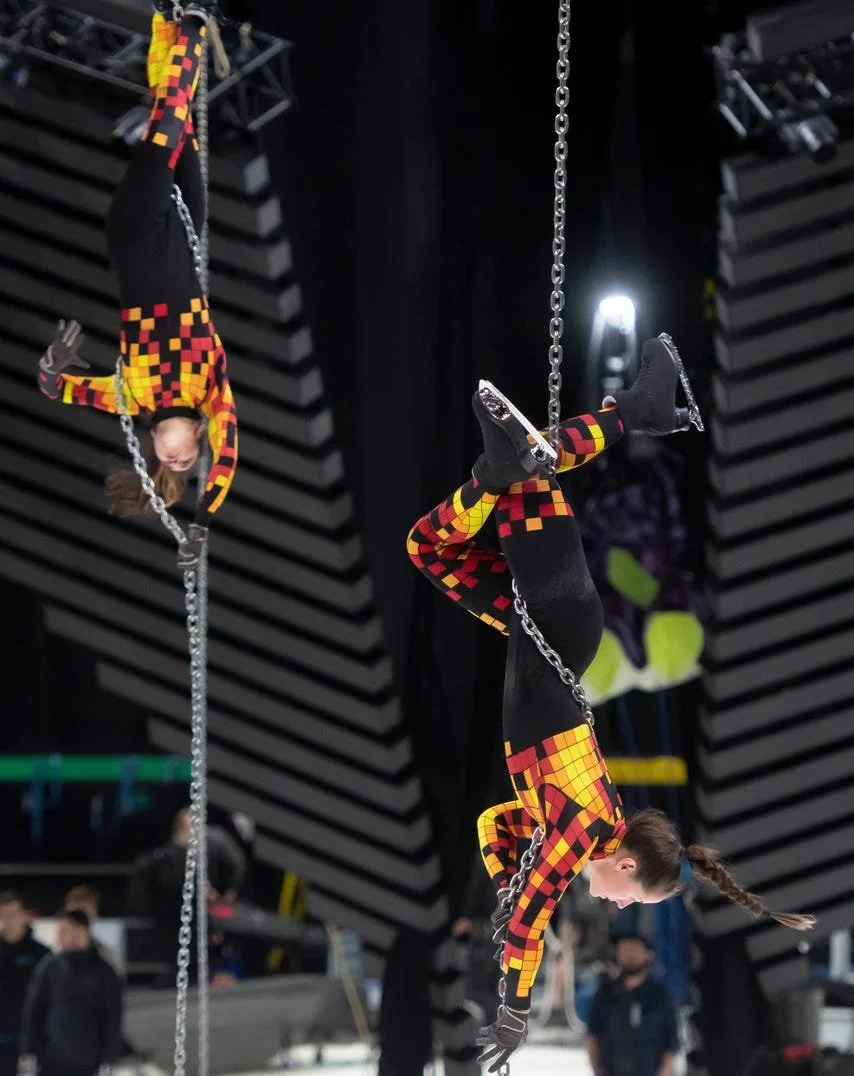 Two performers dressed in black and red checkered costumes hanging upside down on chains in a stage setting.