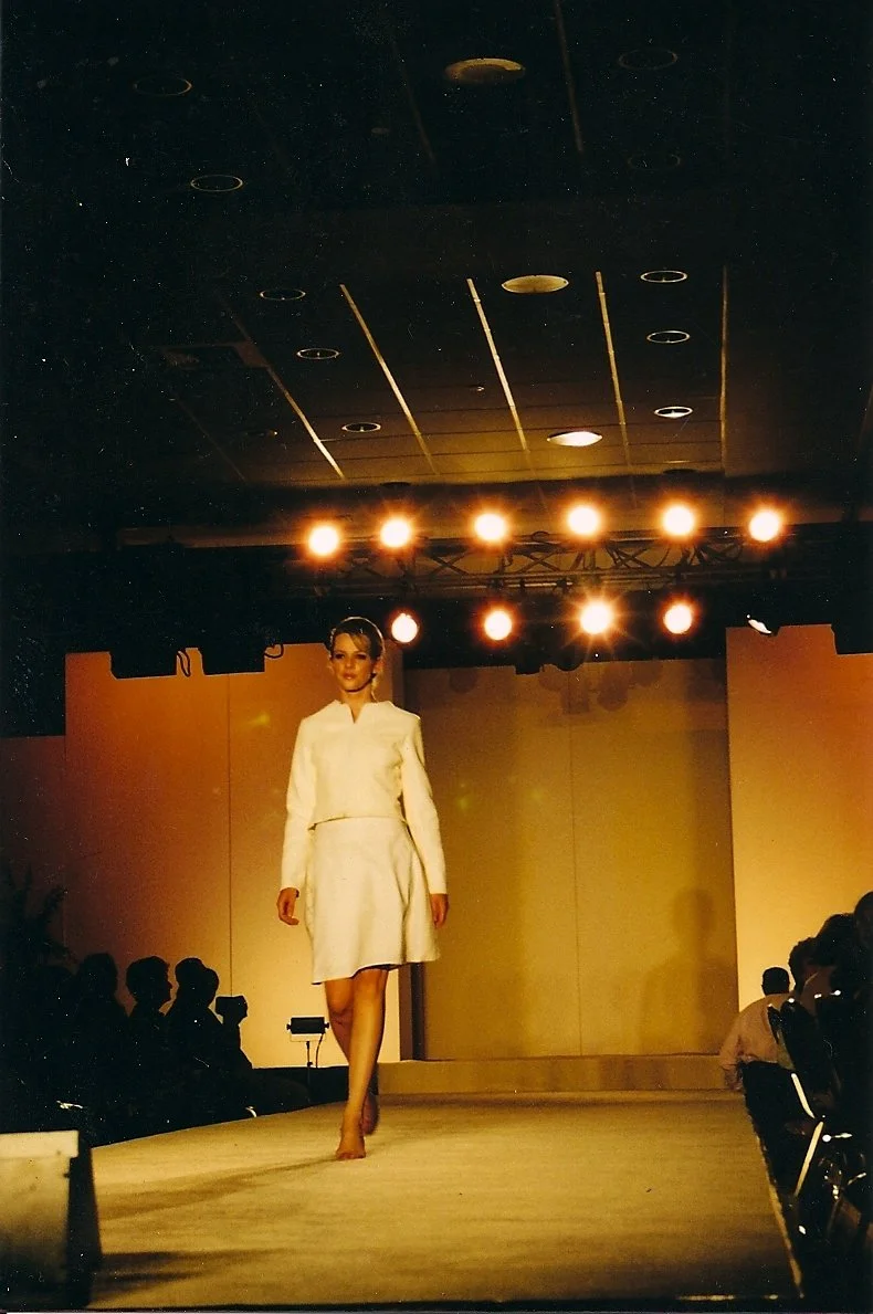 A woman in a white outfit walking down a runway during a fashion show, with the audience seated on both sides and bright stage lights overhead.