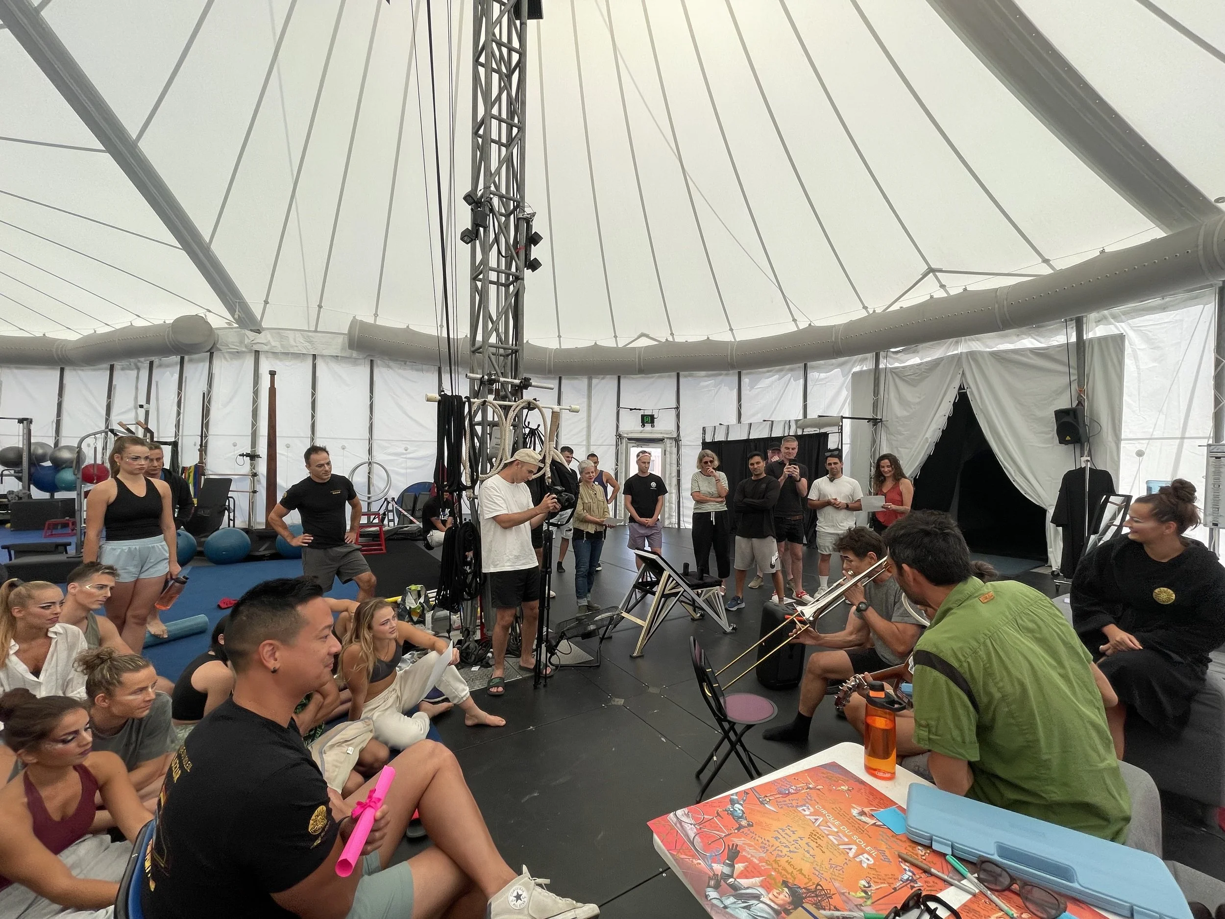 A group of people inside a large, white tent with a high, dome-shaped ceiling. Some are sitting on the floor, while others stand or sit at tables. Several individuals are wearing workout clothes and are gathered near workout equipment, with a few peo
