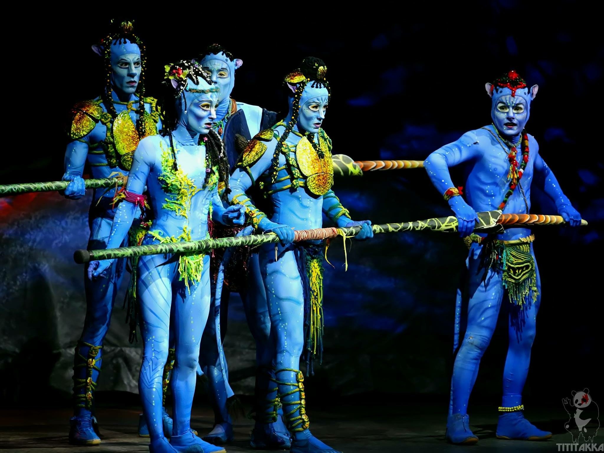 Stage performers dressed as Na'vi characters from Avatar, painted blue with elaborate costumes and makeup, holding a prop spear, with a dark background.