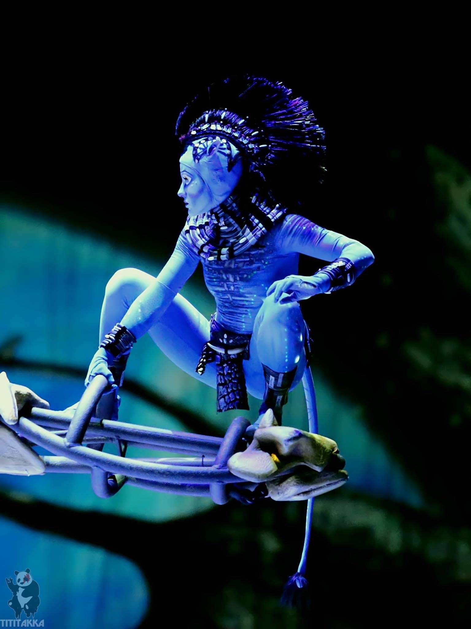 A performer dressed as a character from Avatar, with blue skin and tribal makeup, elaborate headdress, and tribal jewelry, perched on a skeletal structure or bones, illuminated with blue and green lighting.