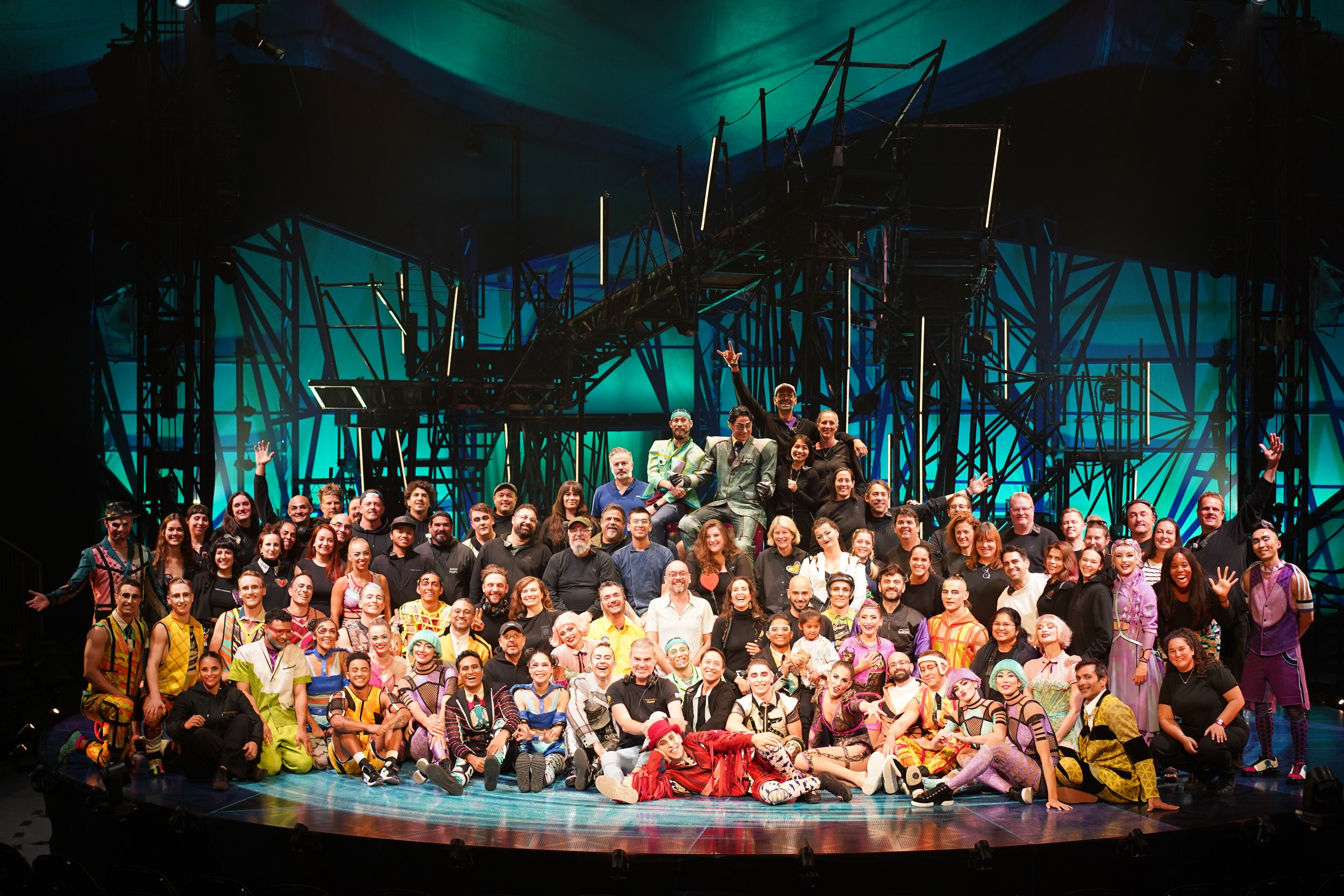Large group of performers and crew on stage after a production of Bazzar by Cirque du Soleil, with colorful costumes and a dark abstract set design in the background.