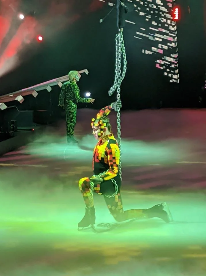 A performer dressed in colorful, pixelated-style costume and makeup, kneeling on one knee on ice skates, holding onto a chain hanging from the ceiling of an ice skating rink. The scene is illuminated with dramatic green and red lighting, with a perso
