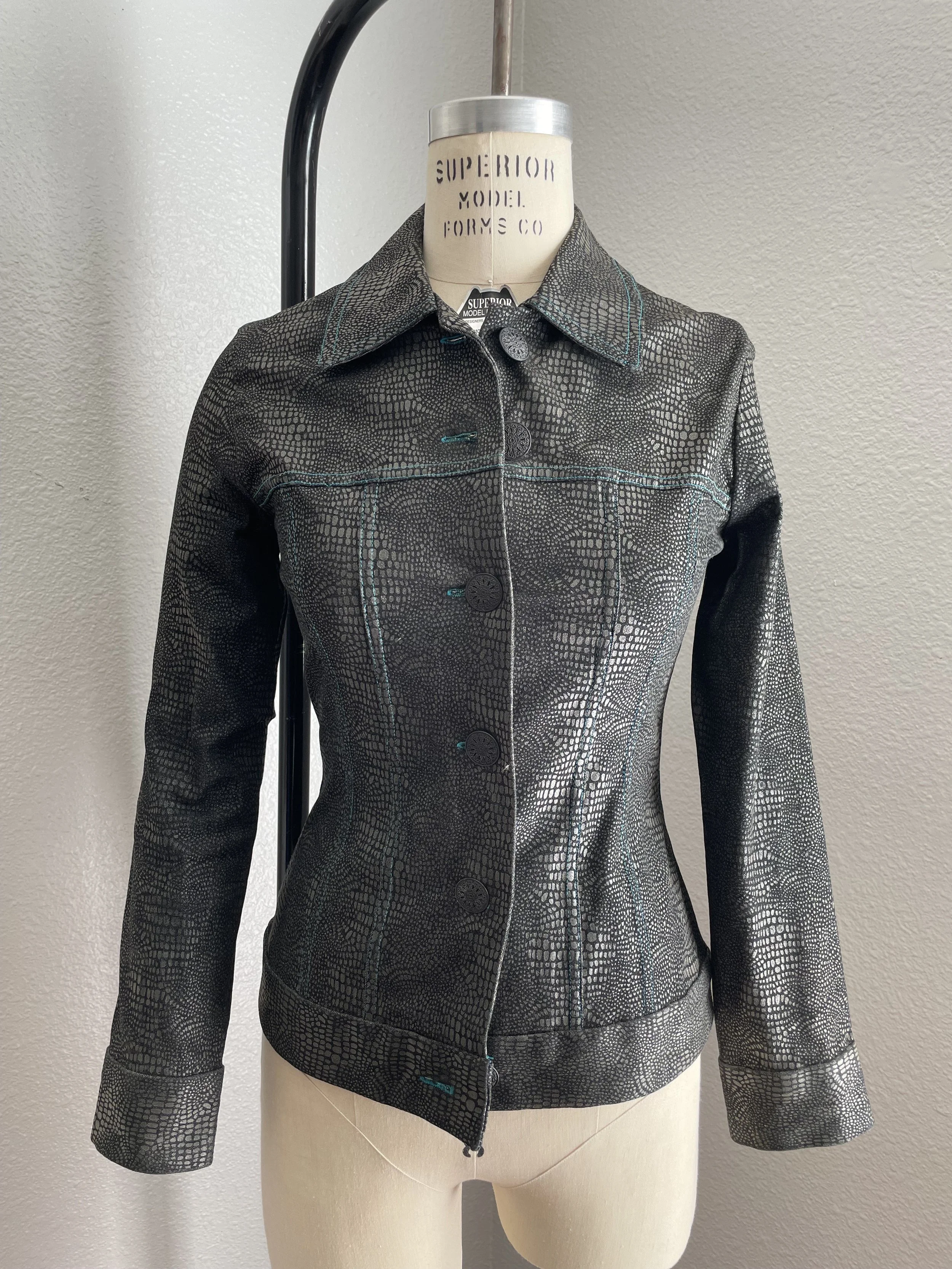 Black and gray textured jacket with teal stitching displayed on a mannequin, featuring a pointed collar and button-up front.