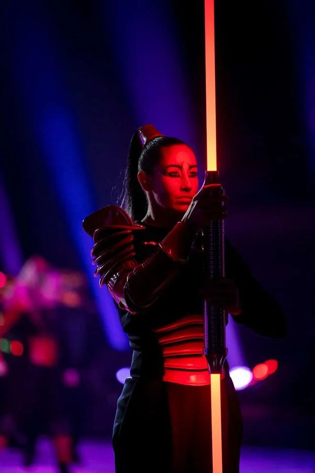 A woman dressed in a futuristic costume holding a glowing red-orange staff in a dark setting with purple and blue lighting.