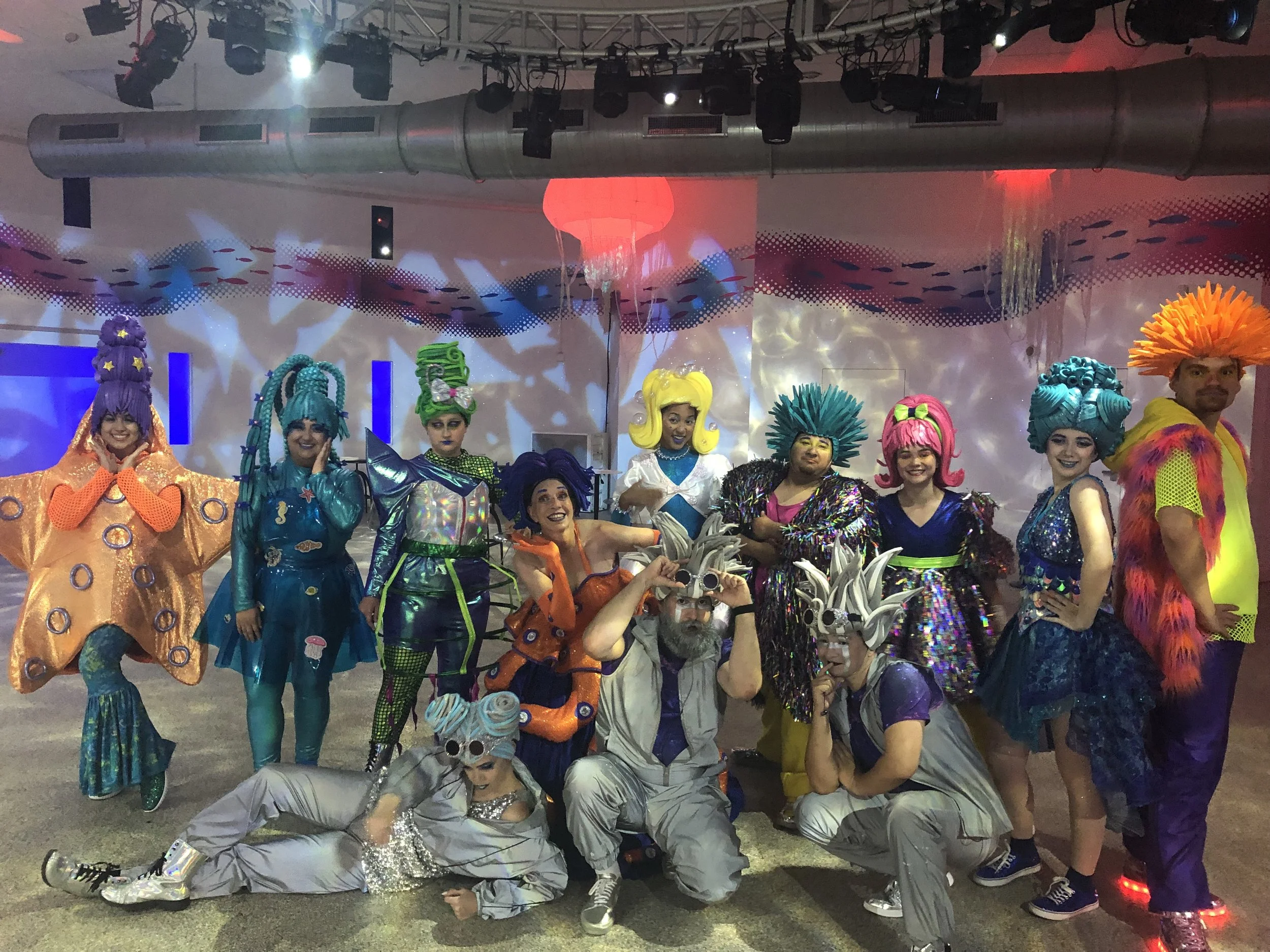 A group of people dressed in colorful sci-fi and fantasy costumes, posing for a photo in an indoor setting with stage lighting and industrial ducts overhead.