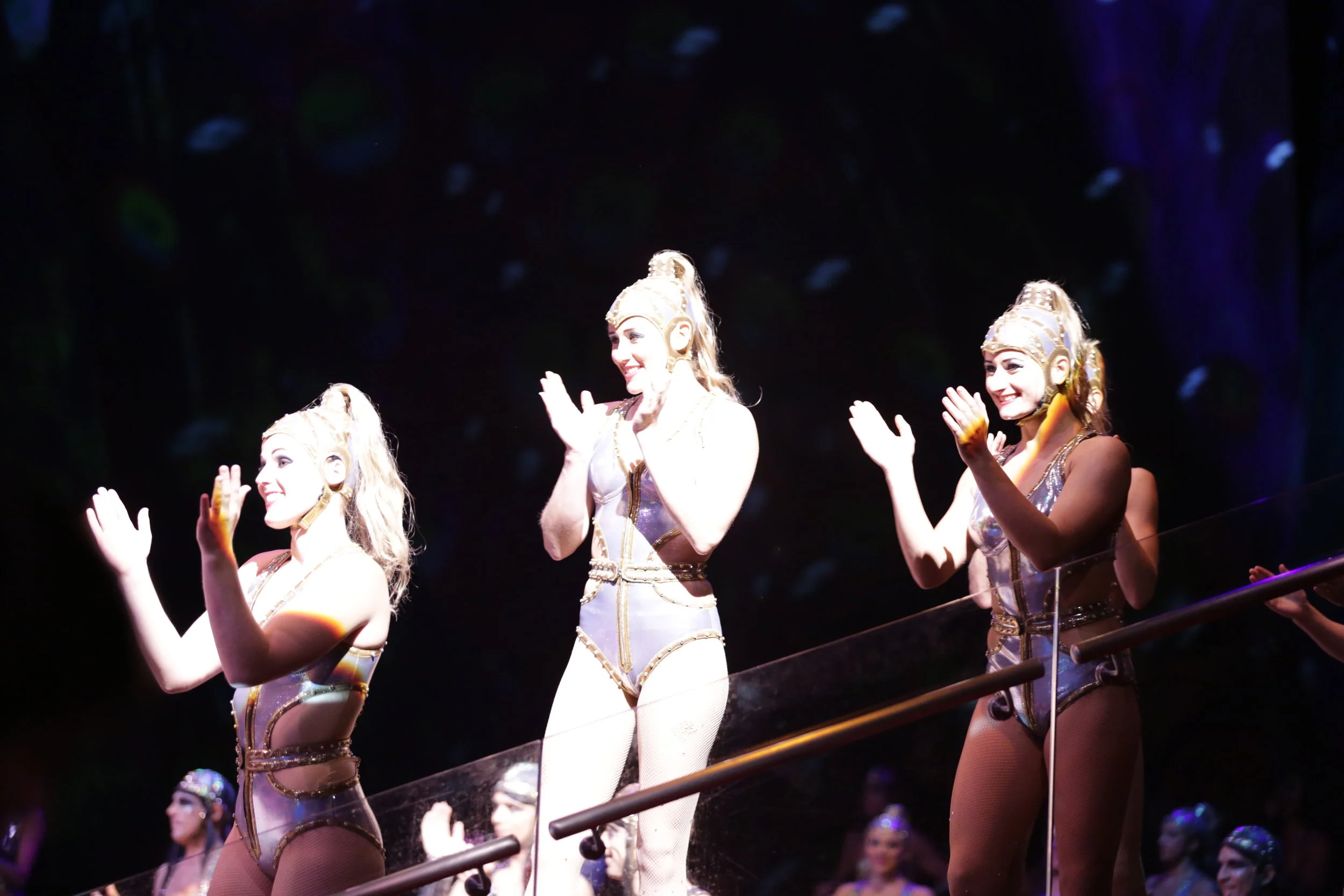 Three performers in elaborate gold and silver costumes clapping on a stage with a dark background and an audience behind a glass barrier.